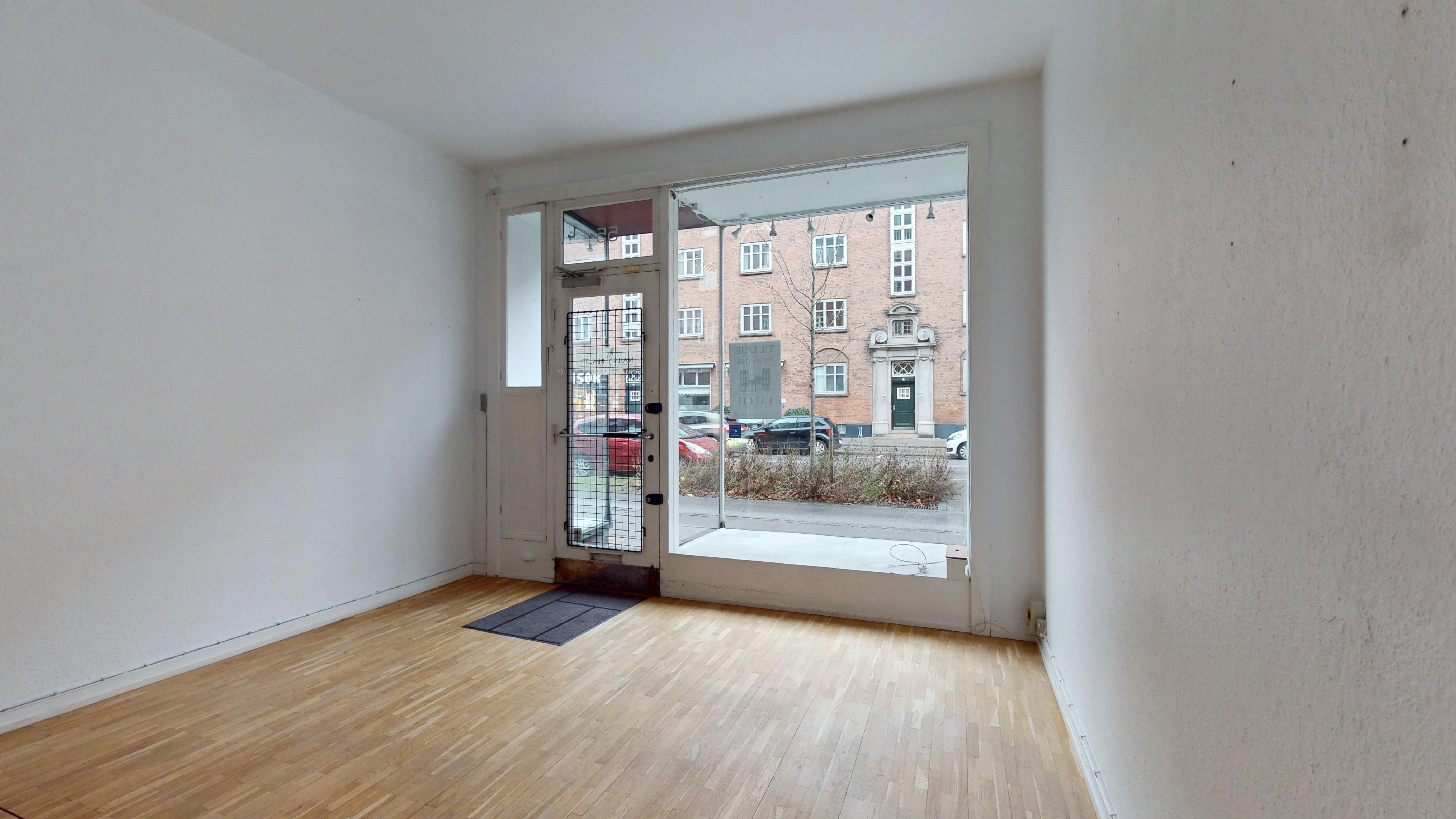 En ren og moderne butik med store ruder og trægulv. En lille indgangsmat og lysekroner i loftet. Bag ruder er der parkerede biler og en bygning.