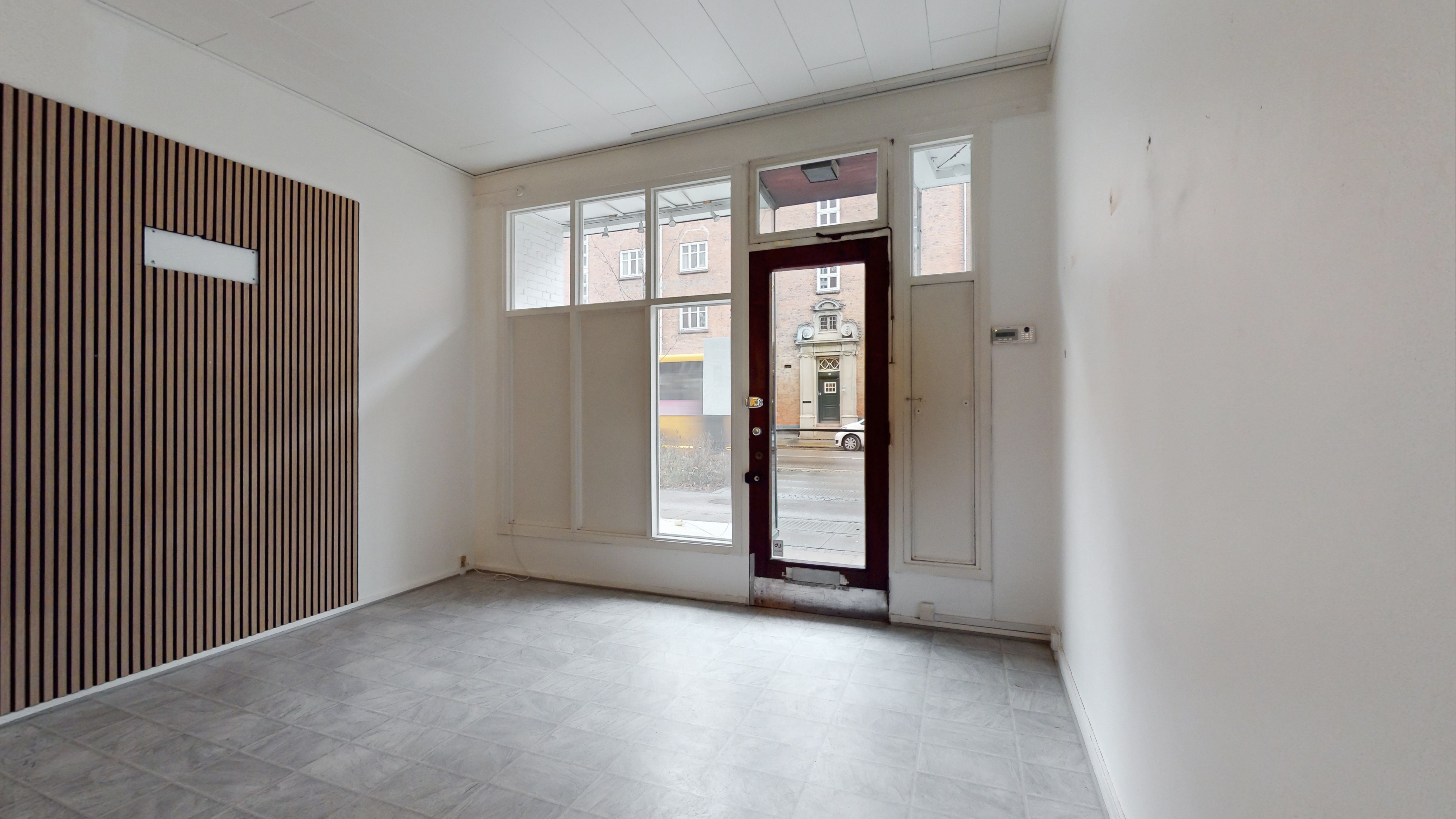 En tom og vellystnet butikslokal med glasruder og træpaneler i gul og grå. Døren er åben, og der er en tydelig udsigt til en gågade med biler