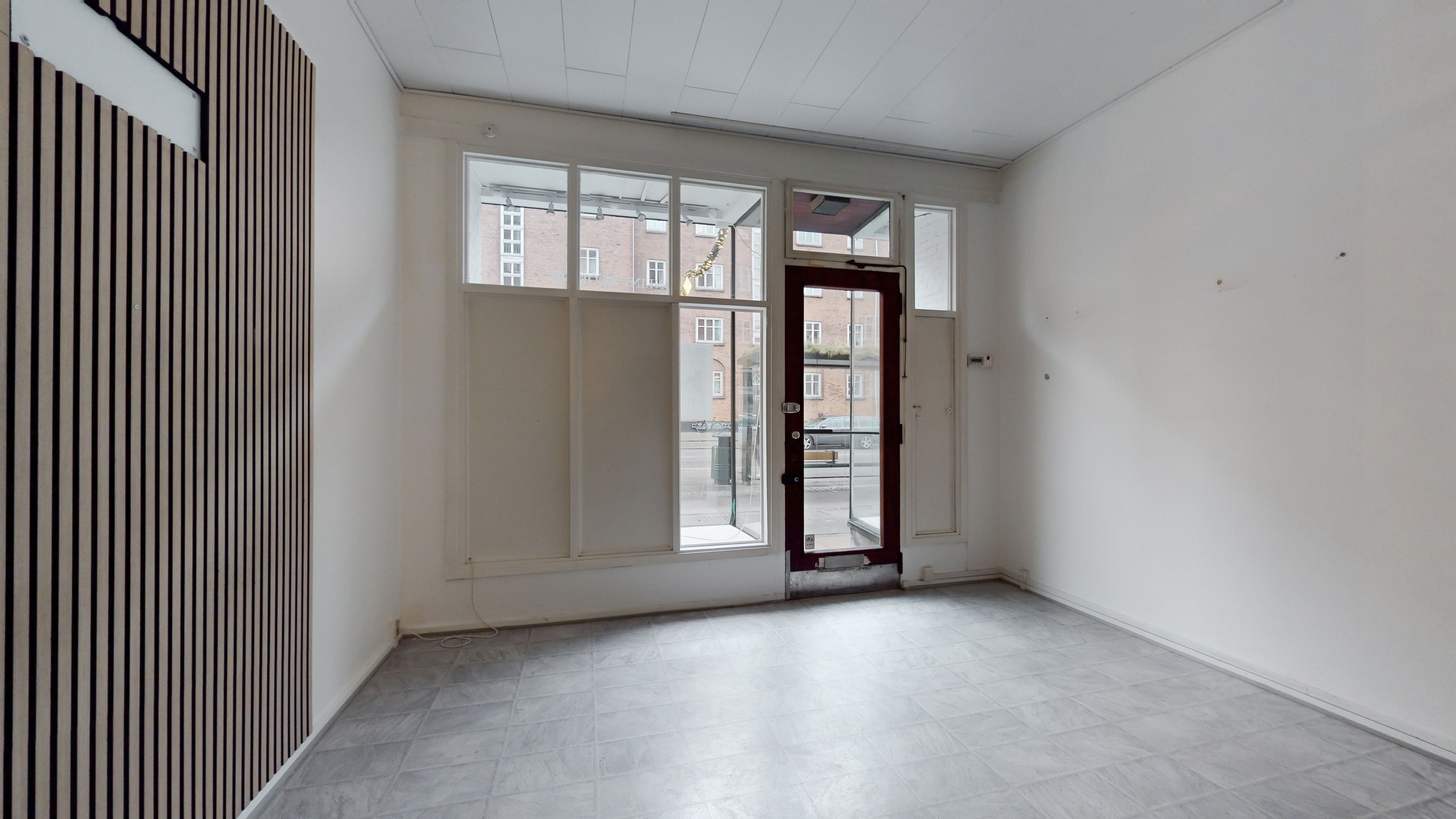 Lukket rum med hvidt loft og vægge samt gulvtæppe. Vindue med gardiner og glasdør med skur mod gaden. Træpaneler og elektriske installationer synlige på væggen.