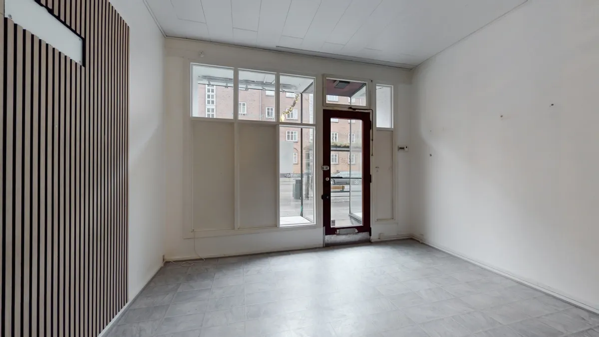 Lukket rum med hvidt loft og vægge samt gulvtæppe. Vindue med gardiner og glasdør med skur mod gaden. Træpaneler og elektriske installationer synlige på væggen.