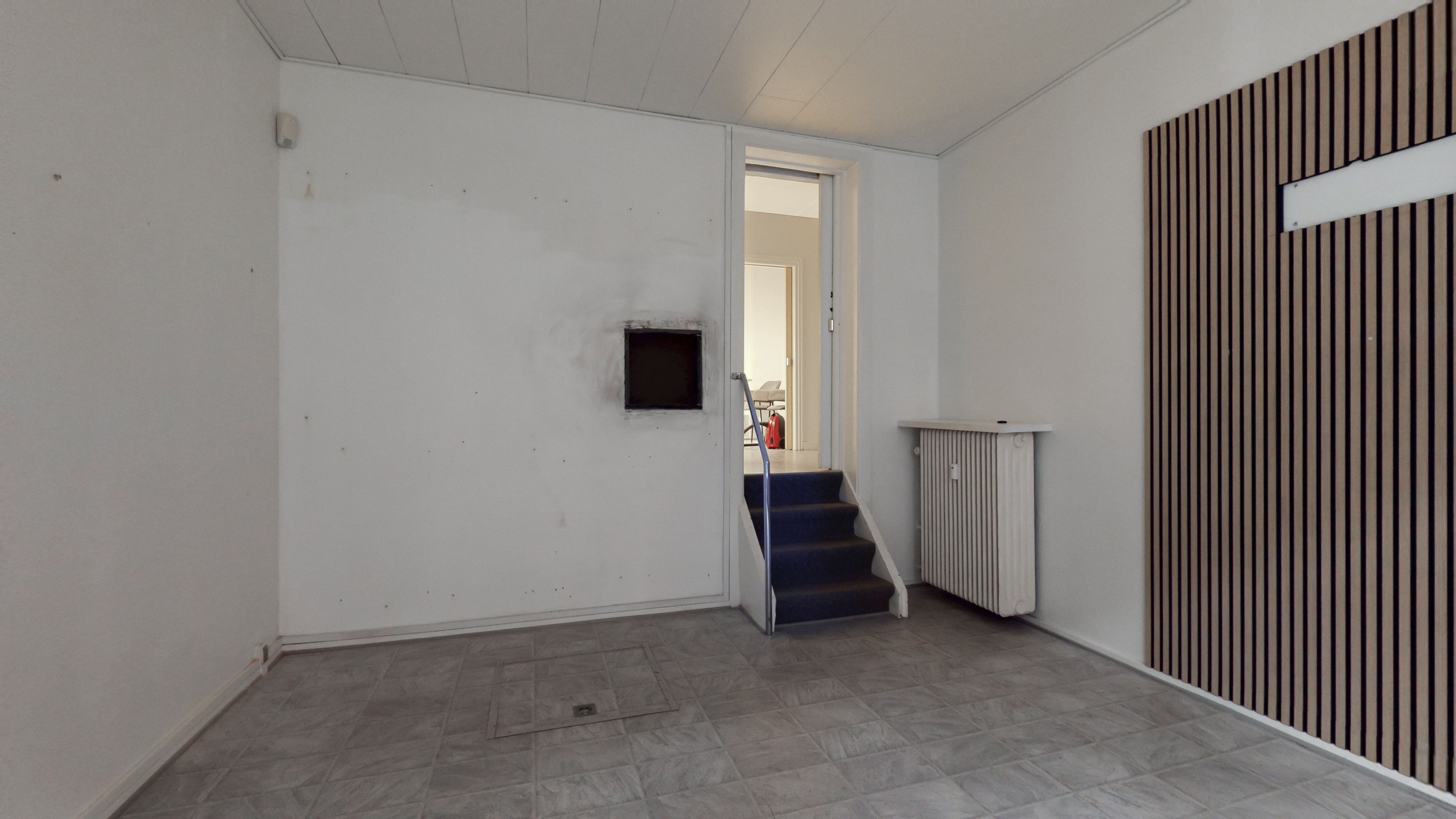 En tom stue med en lysegrå gulvbelægning, en hvid væg og en brun og sort stribet væg med et vindue. Stigende trapper og en radiator er også synlige