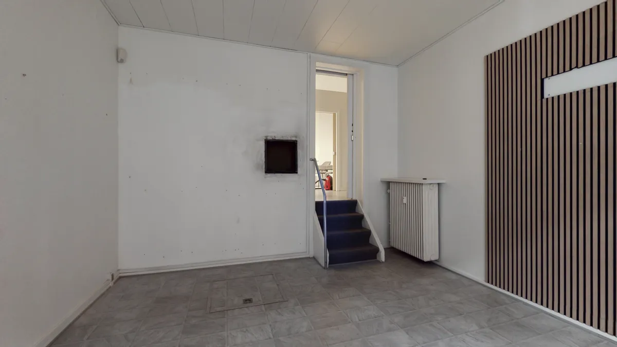 En tom stue med en lysegrå gulvbelægning, en hvid væg og en brun og sort stribet væg med et vindue. Stigende trapper og en radiator er også synlige