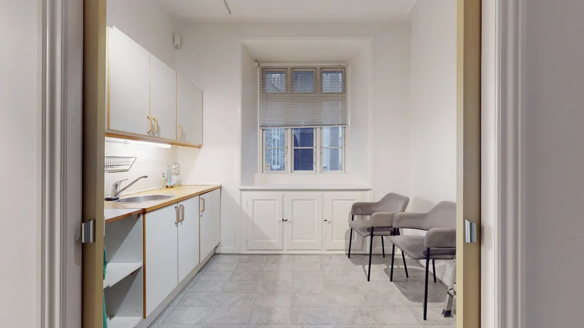 Rent kontor med hvidt interiør, møbler og gulv og to stole til kunderne samt et vindue med jalousier og lys.