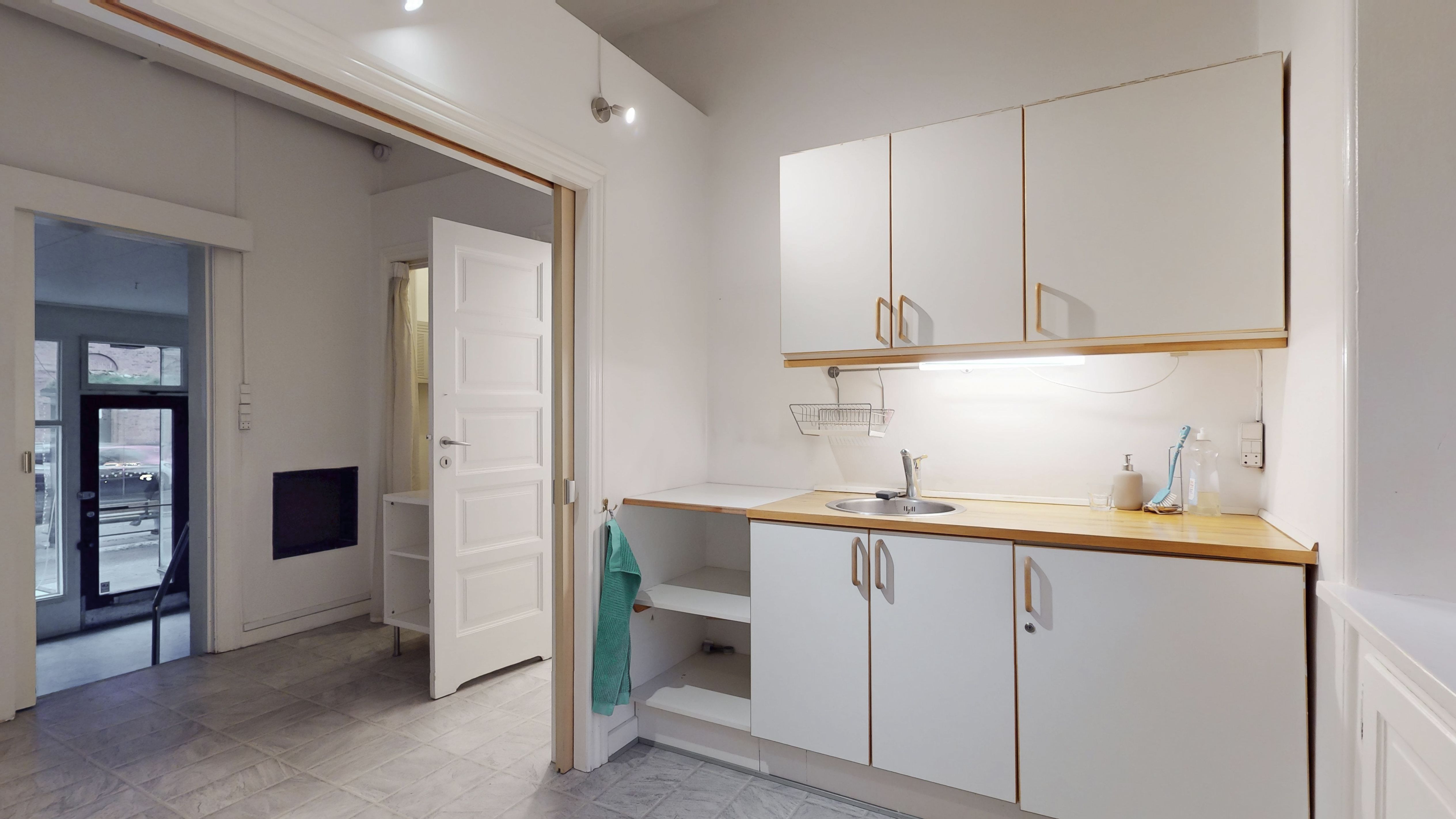 Hvidt køkken med åbne højskabe, lyse gulvfliser og lysegrå vægge samt en åben dør, der fører til et andet rum med en hængekøje