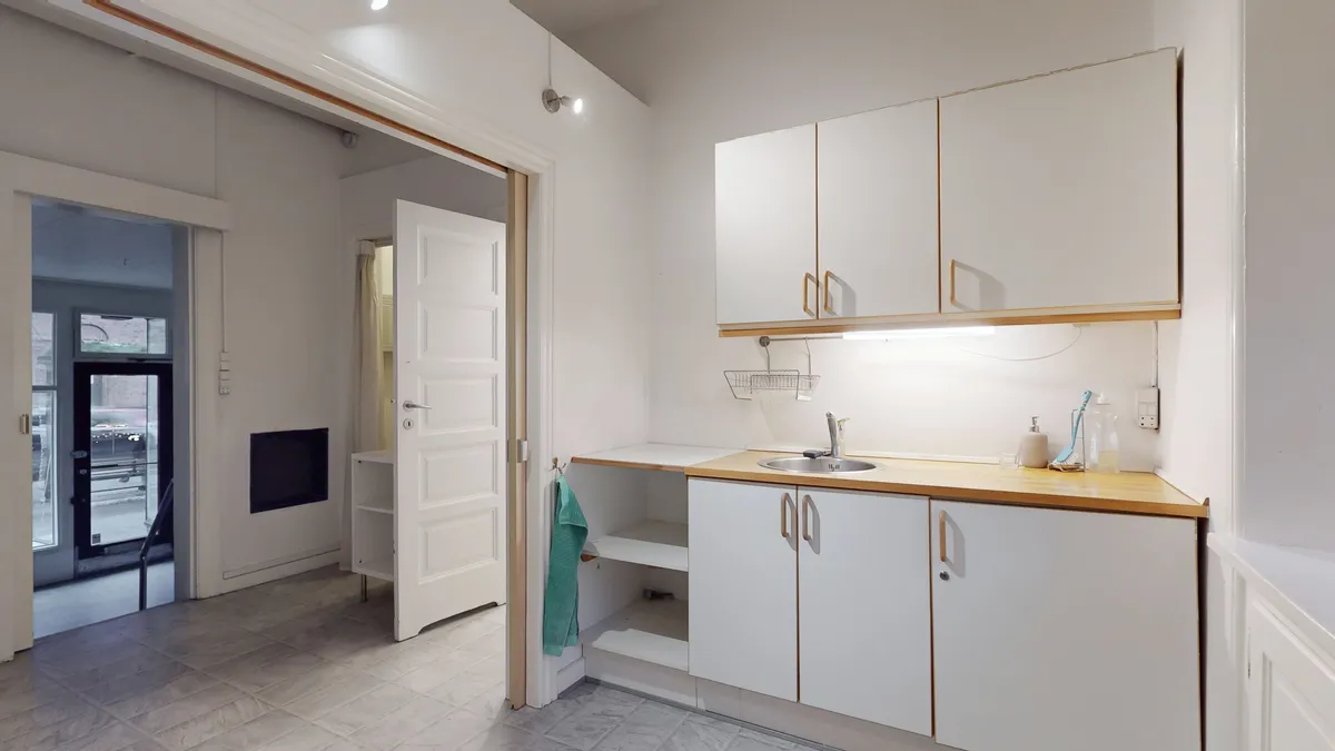 Hvidt køkken med åbne højskabe, lyse gulvfliser og lysegrå vægge samt en åben dør, der fører til et andet rum med en hængekøje