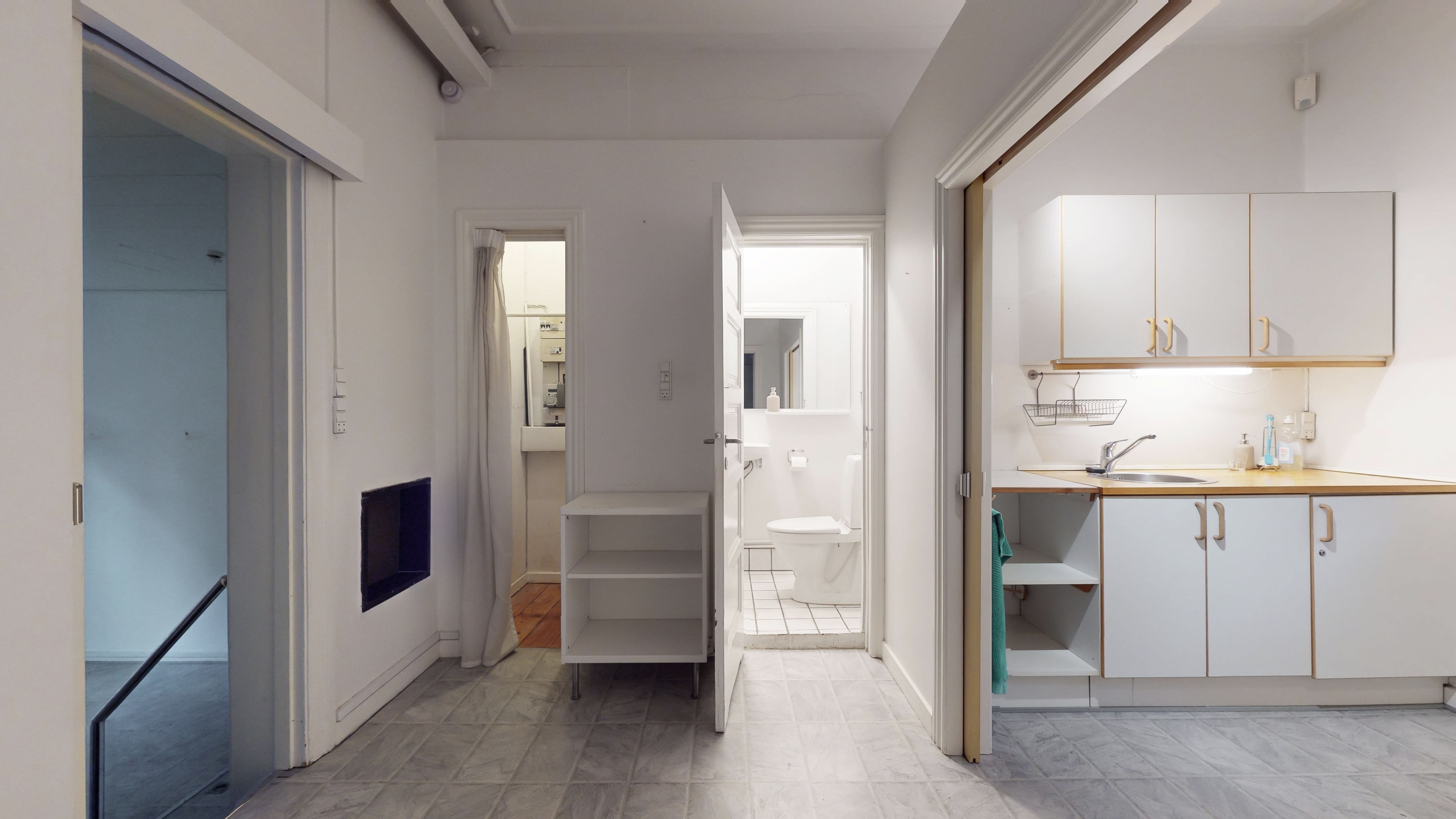 Forretningslejemål med moderne køkken, toilet, håndvask og lyst. Minimalistisk design med hvide vægge og loft. Møbler og håndklæder er også synlige.