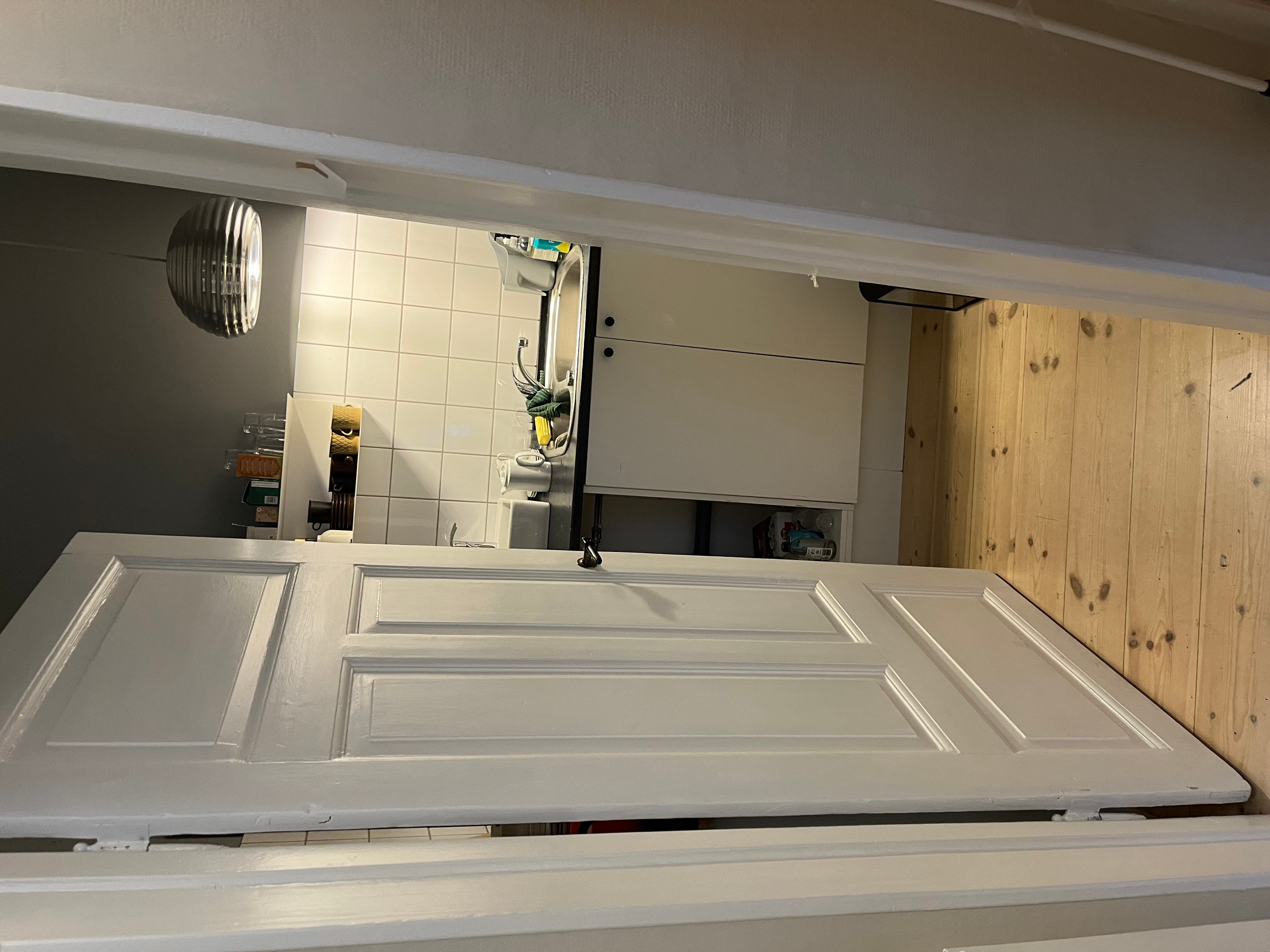 Hjørnekøkken med hvide skabsfronter og lysegrå væg. Sort tørretumbler og lysarm på loftet. Køkken med hvidt loft og gulv.