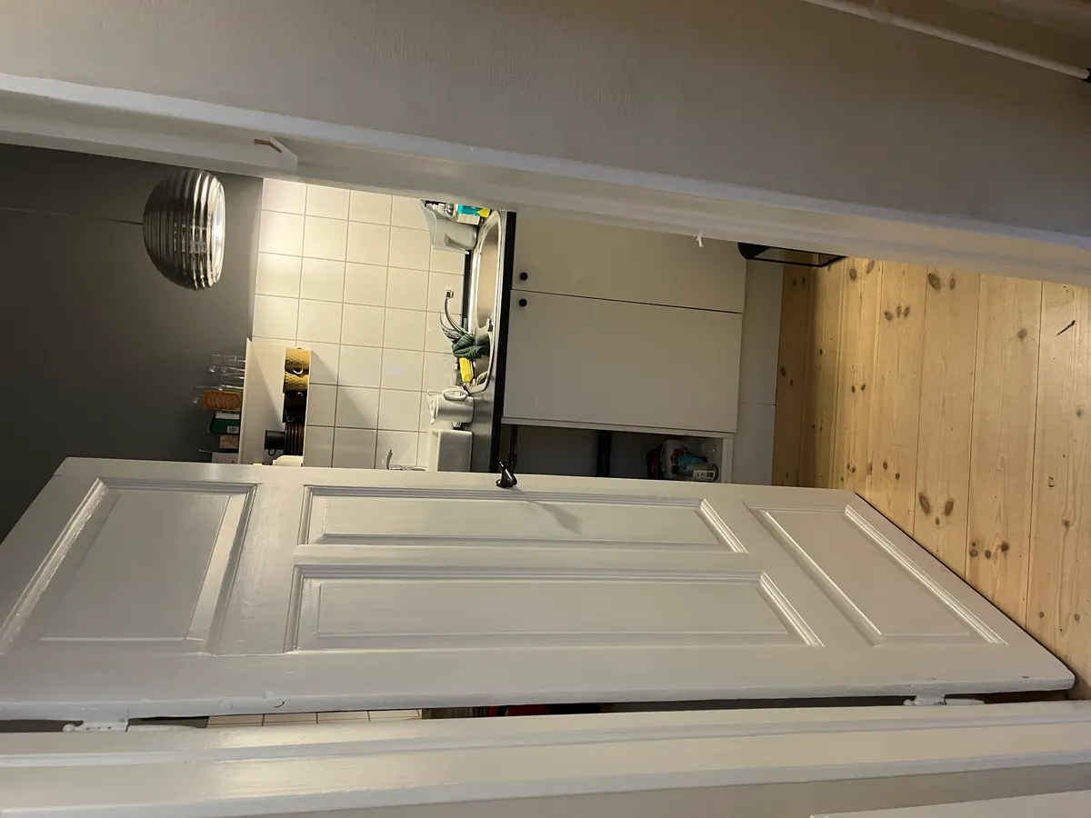 Hjørnekøkken med hvide skabsfronter og lysegrå væg. Sort tørretumbler og lysarm på loftet. Køkken med hvidt loft og gulv.