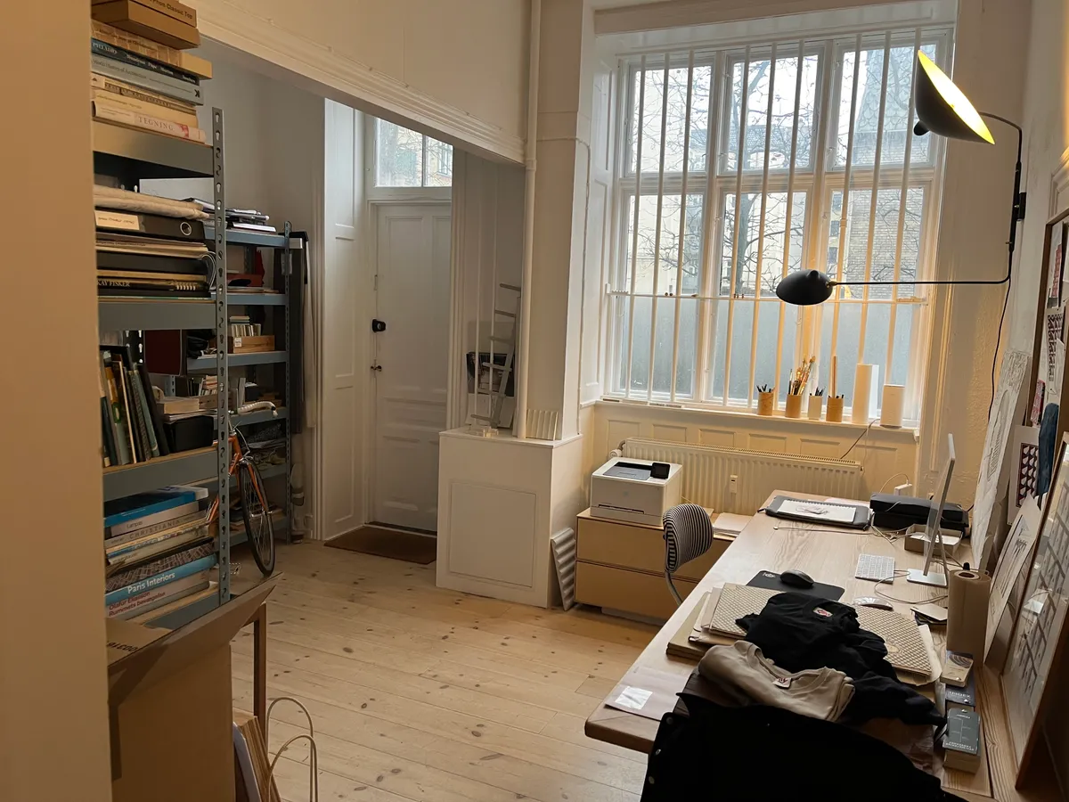 Hyggeligt kontor med skrivebord, computer og lampe. Bibliotek med bøger og høj hylde. Vindue med dobbelte ruder og udsigt til gade. Hvid dør og møbler.