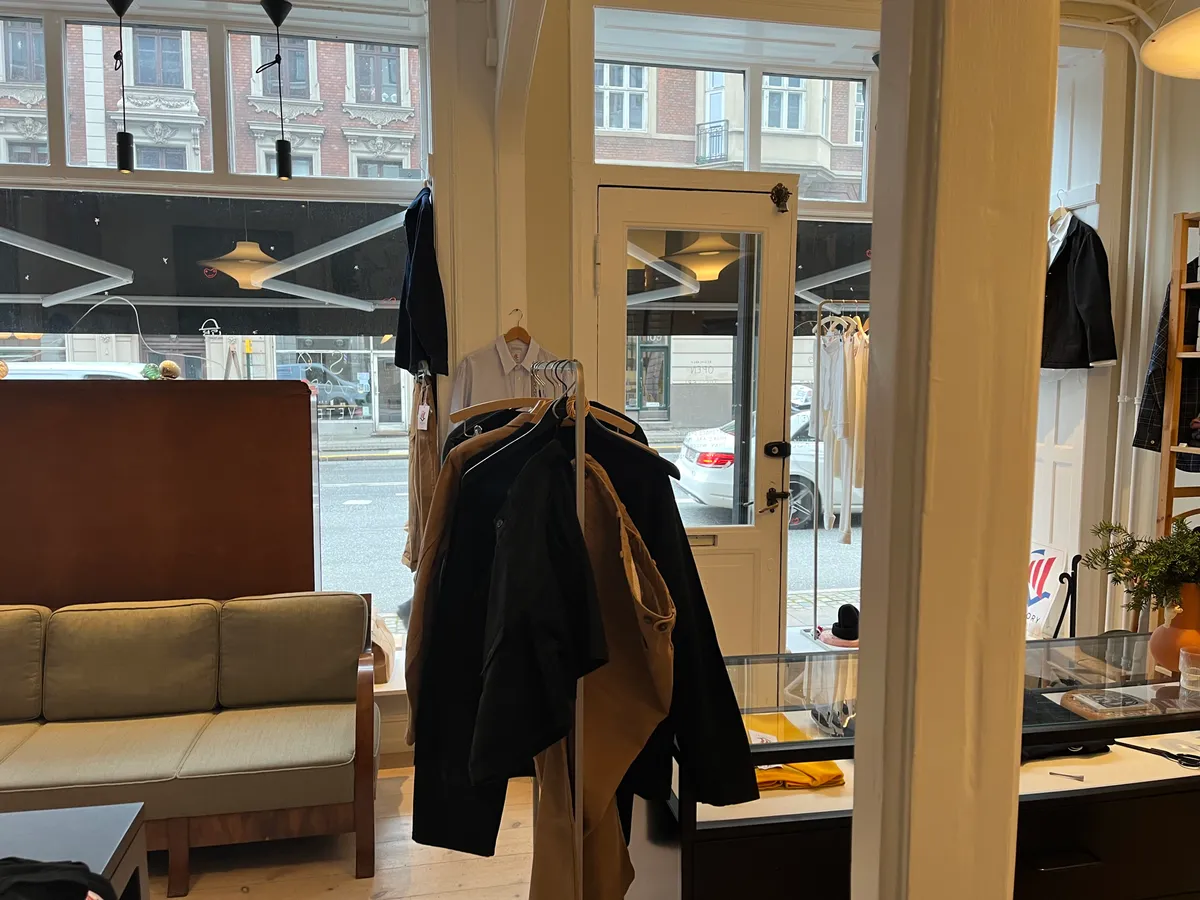 Butikslokale med lyse vinduer og hængende tøj i Frederiksberg, udsigt til parkerede biler og kontorfurnér.