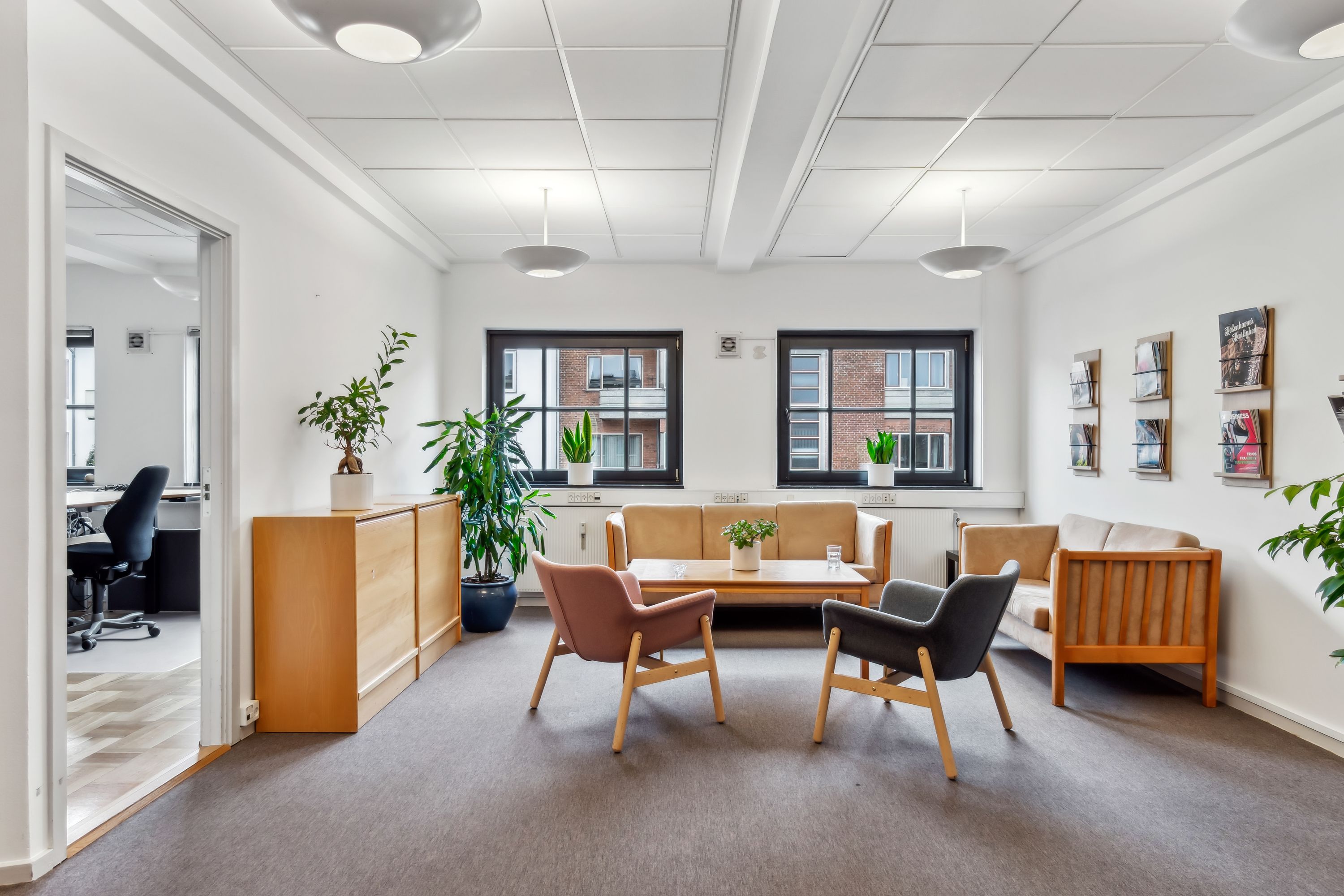 Kontor med moderne møbler, plantegrønne og to vinduer med udsigt til gaden. Et bord med et plantepotte og flere lænestol er placeret på gulvet.
