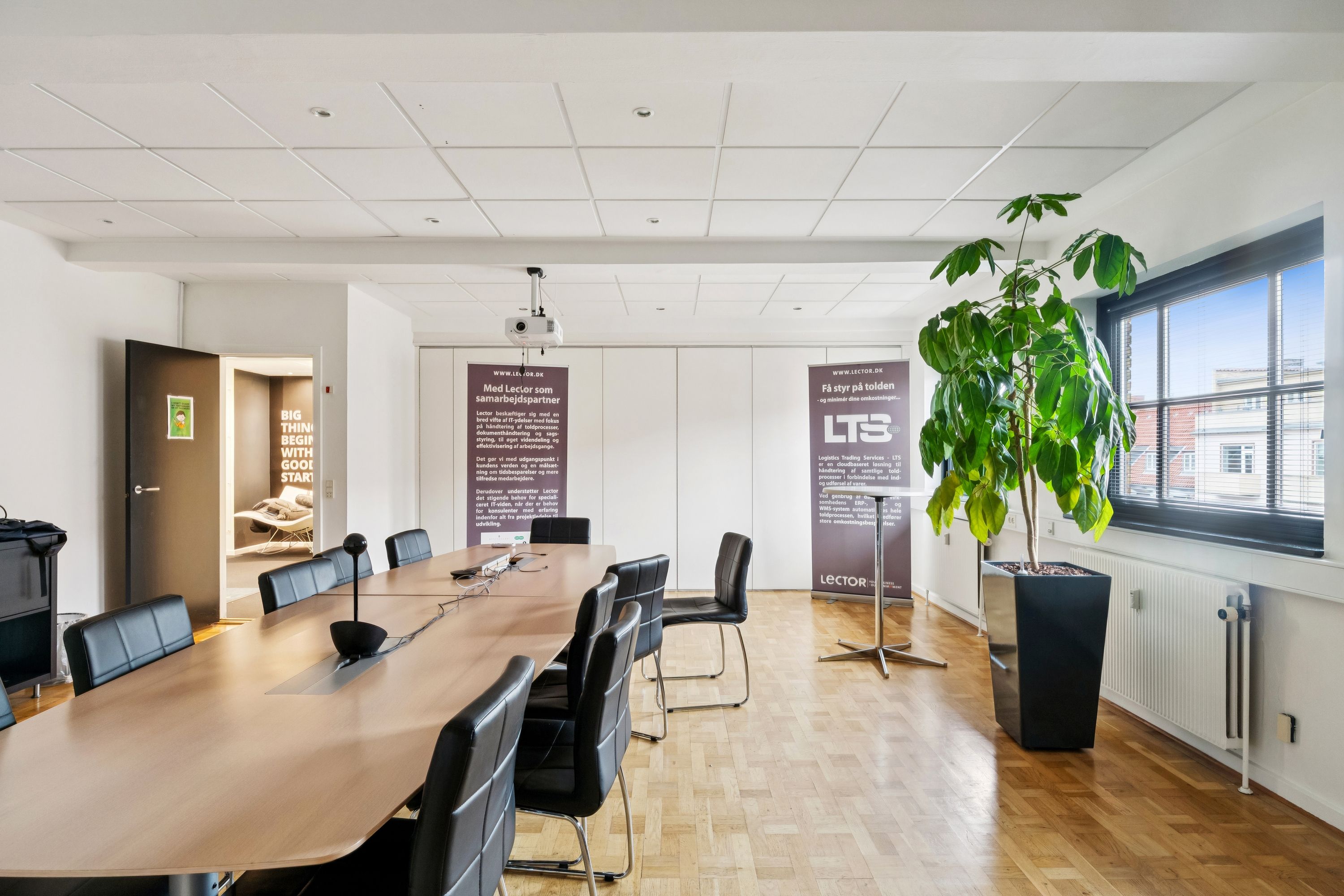 Kontorrum med moderne møbler og faciliteter. En lang mødesal med spidse borde og stole, projektor og radiatorer. Planter og reklamebannere tilføjer naturlige elementer og branding til kontoret.