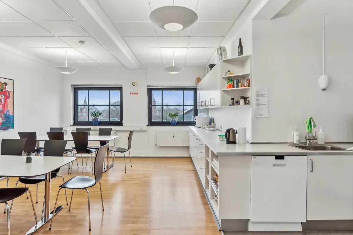 Kontorlokaler med moderne interiør og trægulve, vinduer med udsigt til grønne områder, bord og stole, køkken med moderne indretning, hvide vægge og loftslampe
