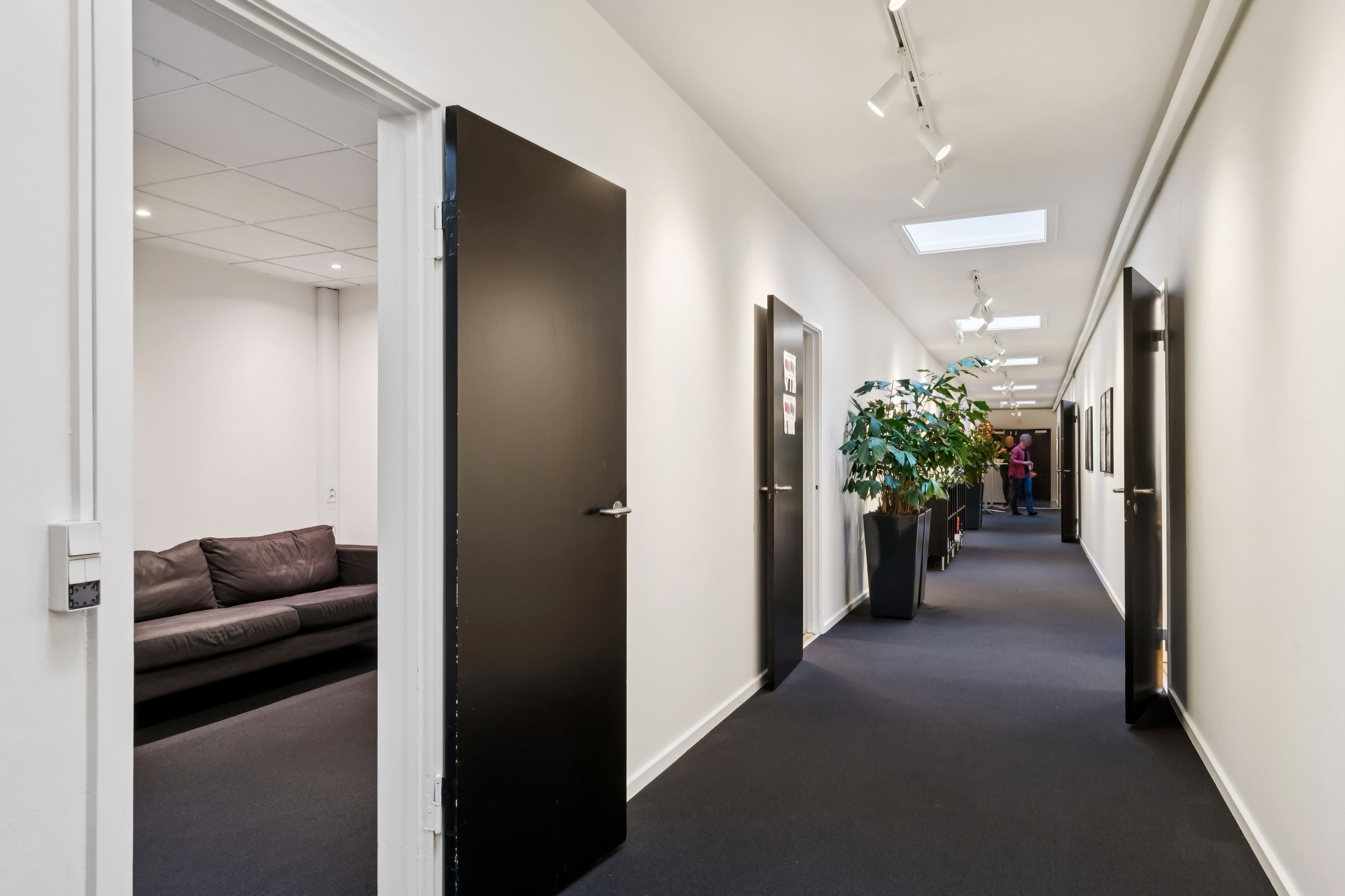 Kontor med indgangsparti, sofa, plante og to elevatorer i hvid og sort. Gulvet er dækket med sort gulvtæppe og der er hvide vægge
