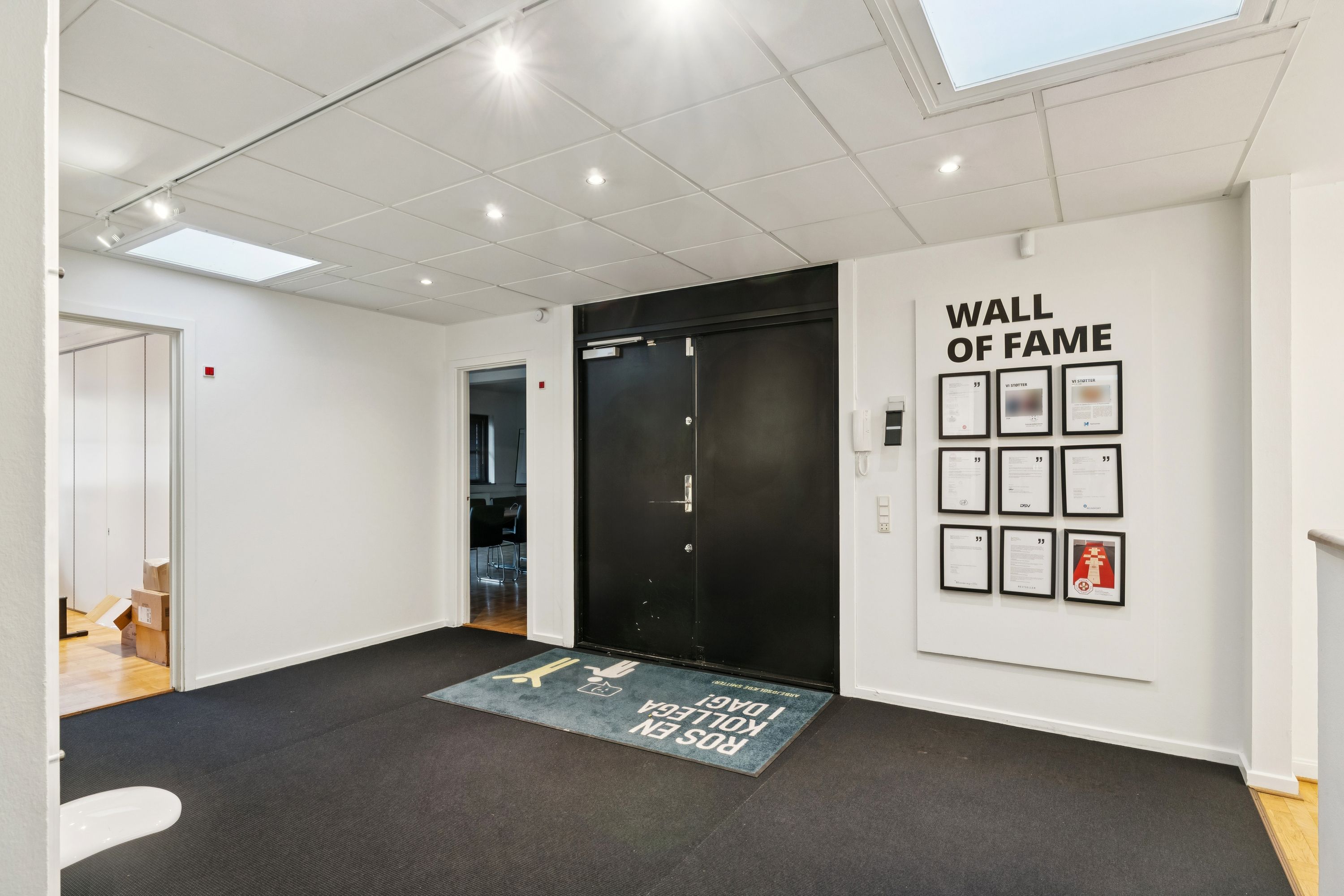 Indvendigt er der hvide vægge, gulvtæppe og lyskryds på loftet. Der er tekst på væggen og gulvtæppet læser Booting Please Wait. Der er en dør med tekst og en væg med Wall of Fame