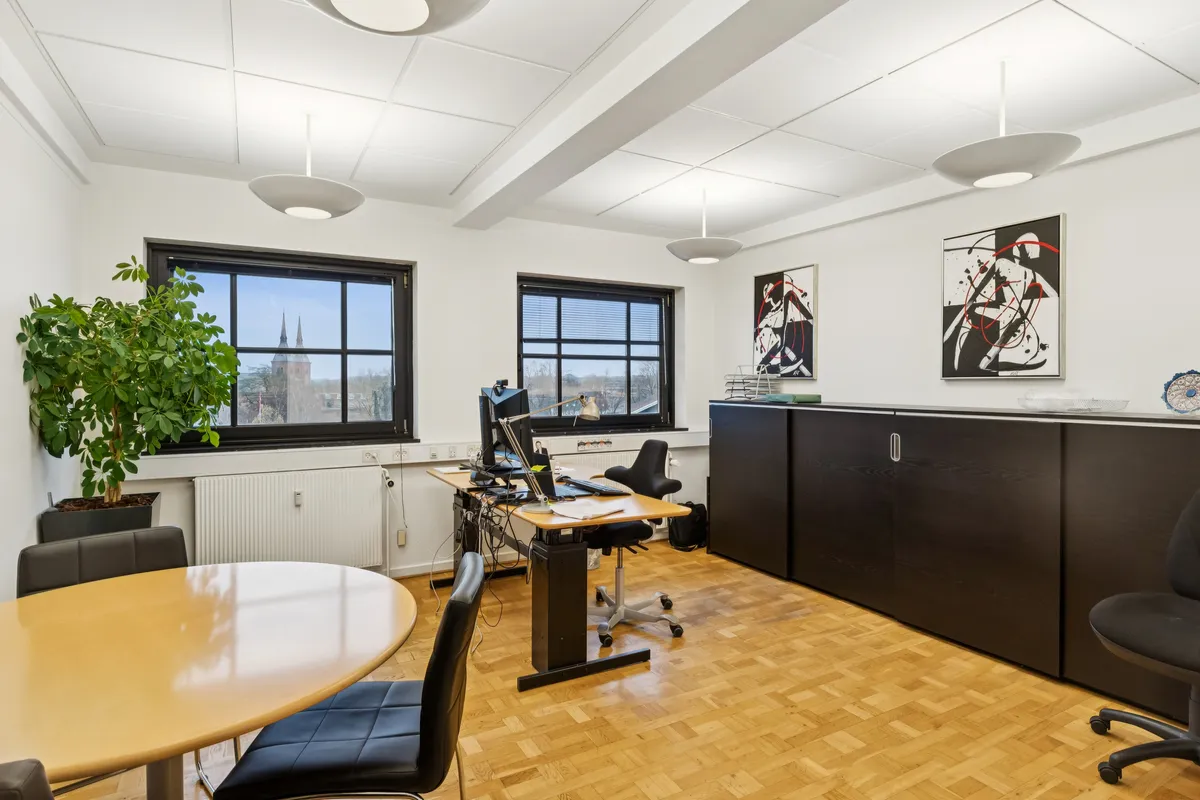 Kontor med to skriveborde, computere og skrivebordslamper samt tre stole. En plante og to malerier er hængt på væggen. To vinduer er åbne