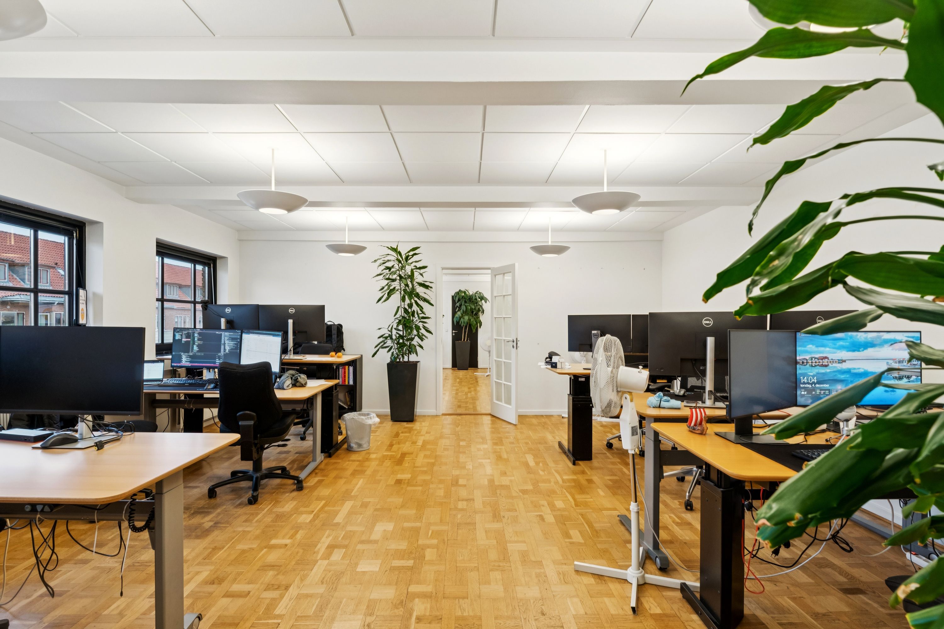 Kontorrum med moderne skriveborde og computere, grønne planter og hvide vægge samt loftslampe. Værelse med åben dør og vinduer, der ud til bygningen.