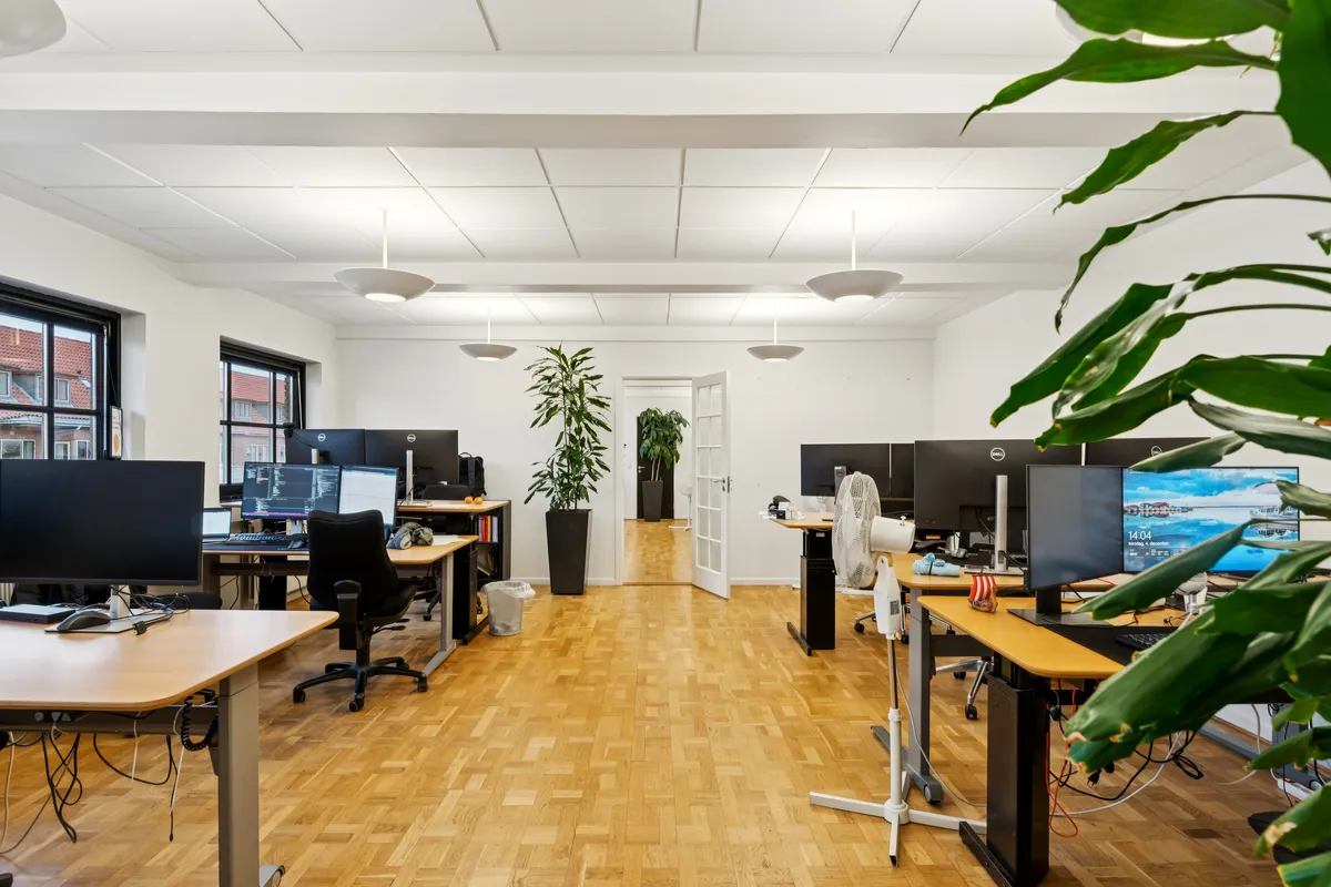 Kontorrum med moderne skriveborde og computere, grønne planter og hvide vægge samt loftslampe. Værelse med åben dør og vinduer, der ud til bygningen.