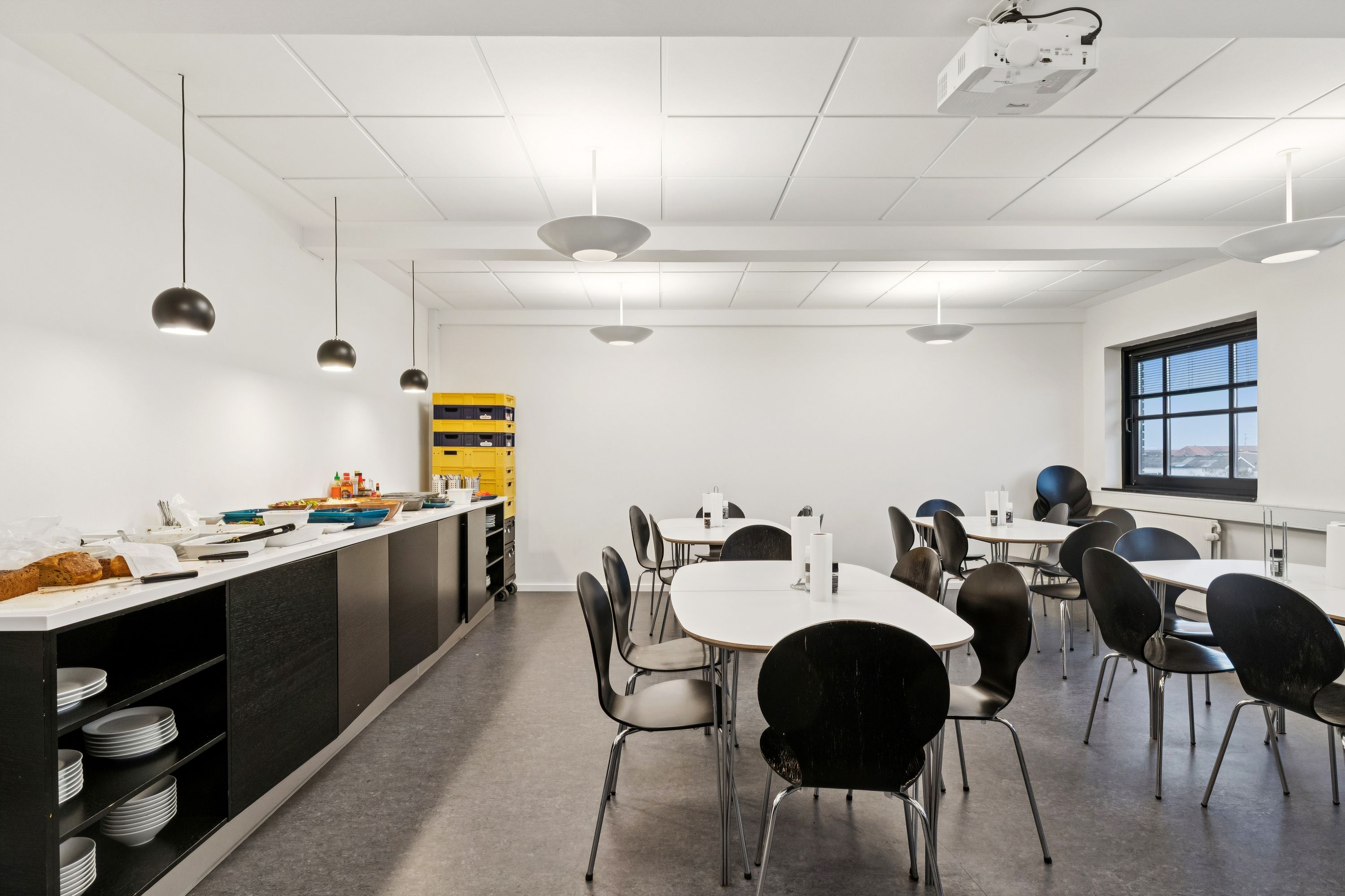 Kontor med moderne indretning og minimalistisk design, hvor borde og stole er placeret og lyskroner er hængt på loftet. Væggene er hvide og der er stolpebord med mad og drikkevarer.