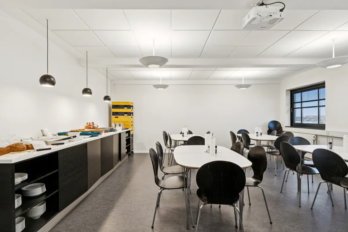 Kontor med moderne indretning og minimalistisk design, hvor borde og stole er placeret og lyskroner er hængt på loftet. Væggene er hvide og der er stolpebord med mad og drikkevarer.