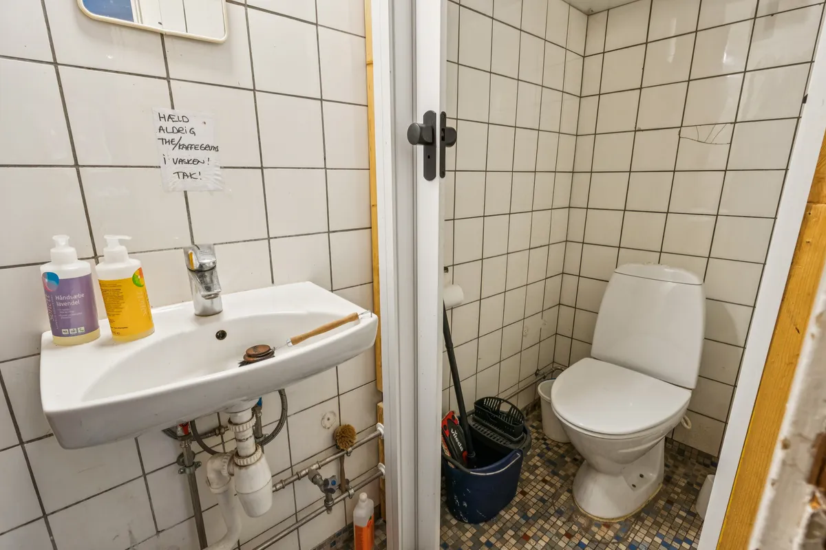 Billede af et lille badeværelse med en hvid toilet, en hvid vask, og en håndsæbeflaske og en sæbeflaske på vasken og et rengøringsværktøj i en blue søppelspande