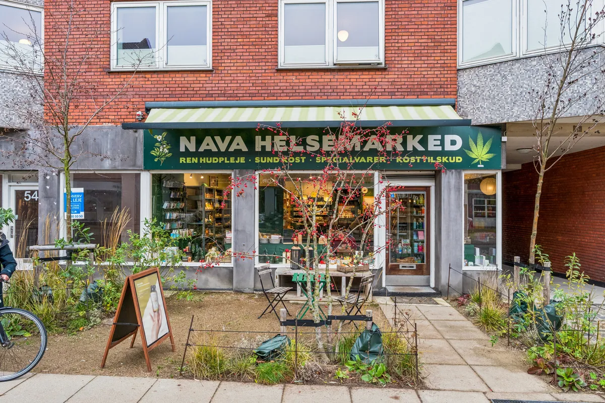 Butik med grønt banner, udsalg af urter og plantebaserede produkter og have med rækværk. Der er en cykel og stol og bord med grønne produkter udenfor butikken.