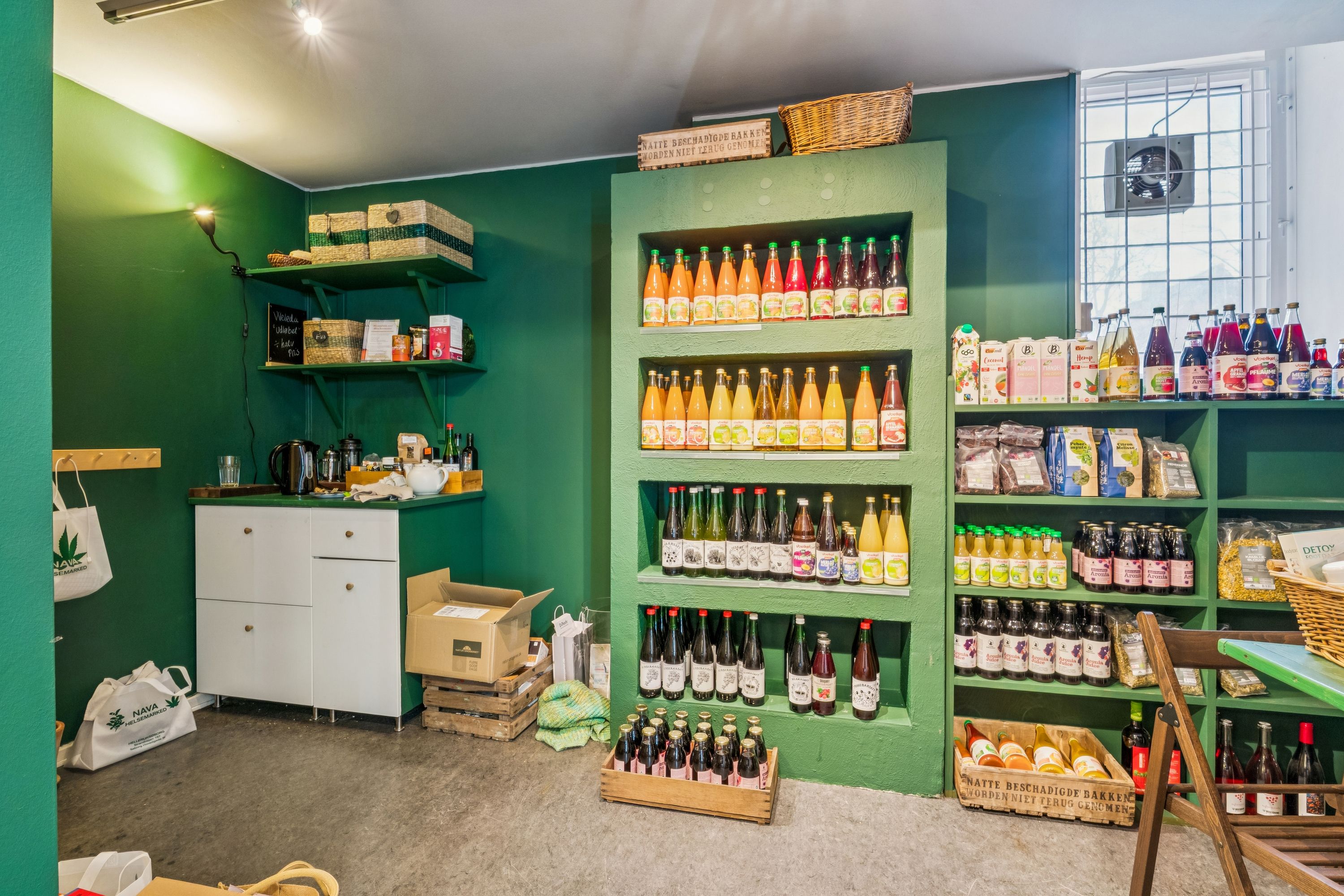Butik med grønne vægge og hylder med diverse drikkevarer og produkter, bl.a. æblemost, kartofler og bagværk, samt en hvid kommode og trækurv på gulvet