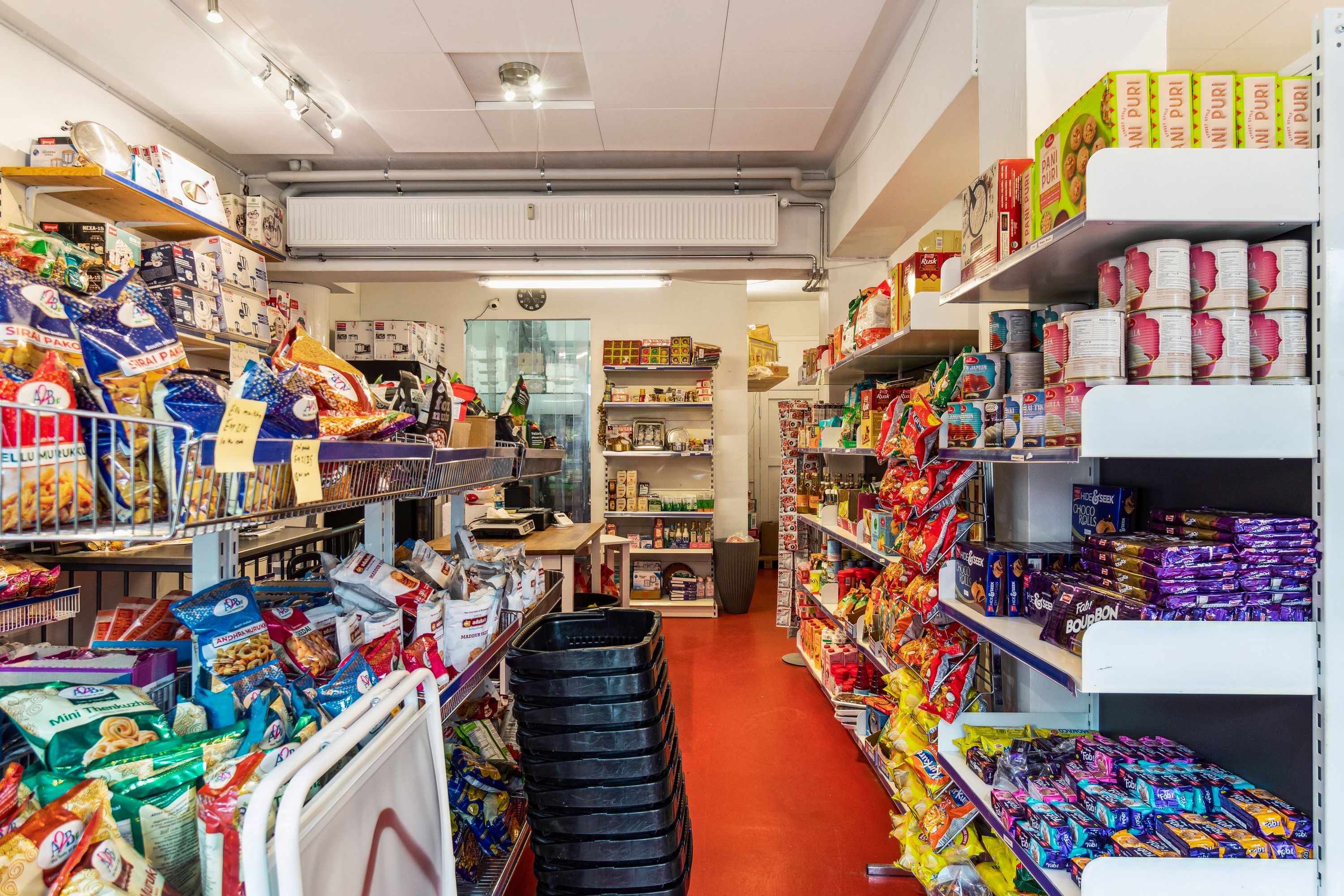 Butik med snacks og chokolade i lager og på hylderne. Sorte kurve og et bord med en vægt. Lyse loftslamper og rødt gulv.