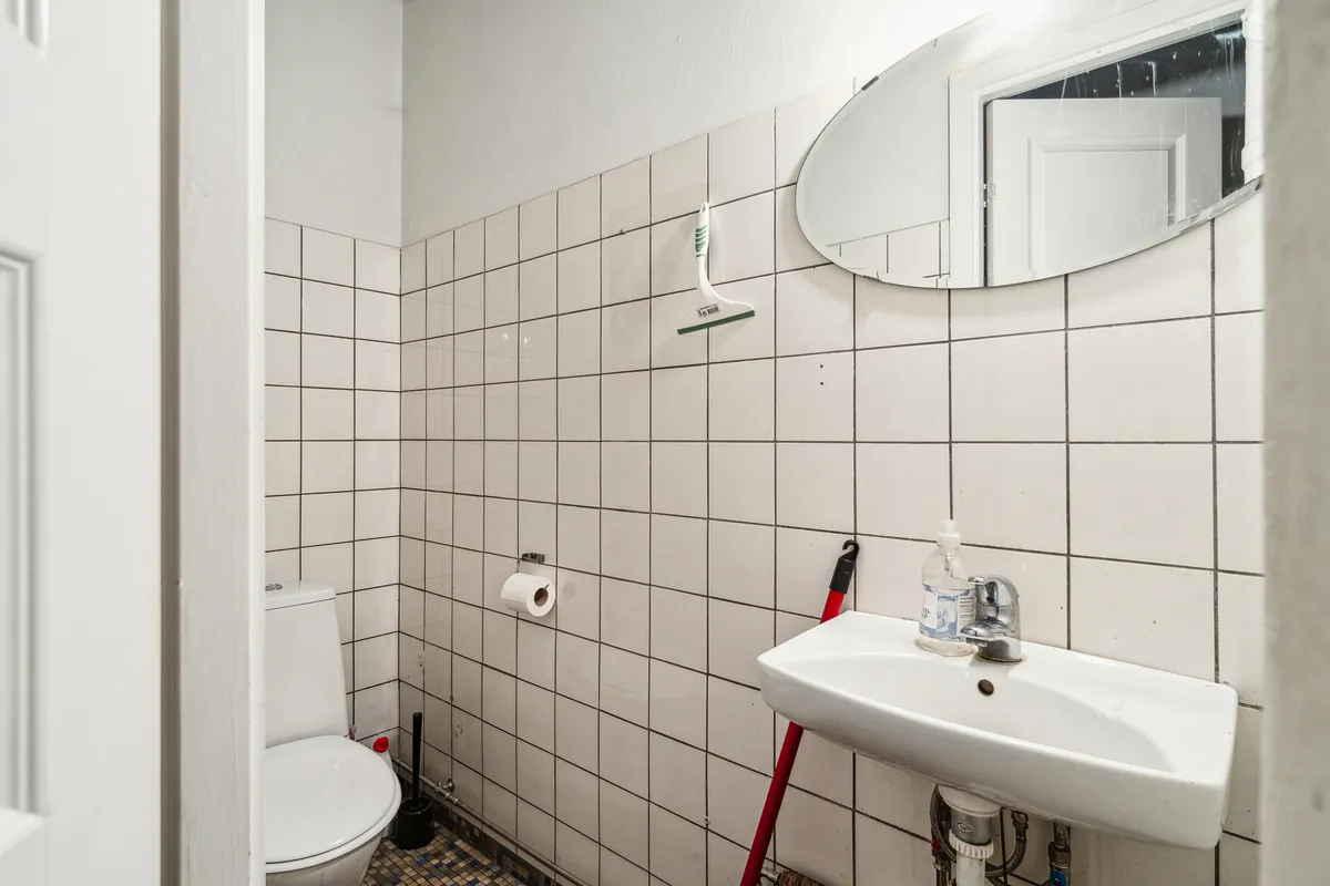 En lille toilet med hvidt toilet, hængende skraber og skraber, toiletpapir og en hvid sænke med vandhane og håndsæbeflaske. De hvide keramiske væggen og gulvet med et spejl i loftet.