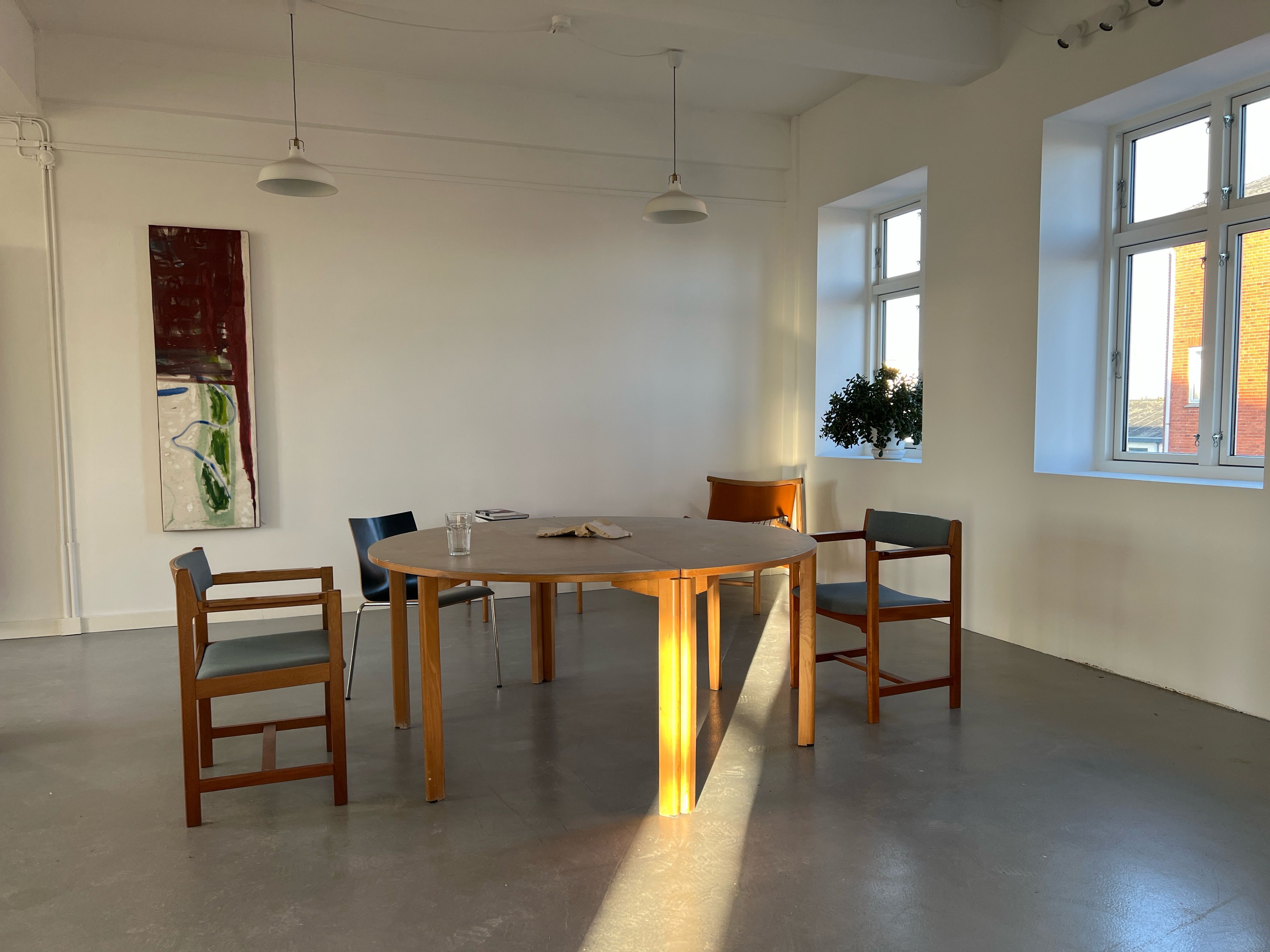 Kontorrum med rundt bord og fire stole samt maleri på væggen og vindue med plante og ududsigt til bygning