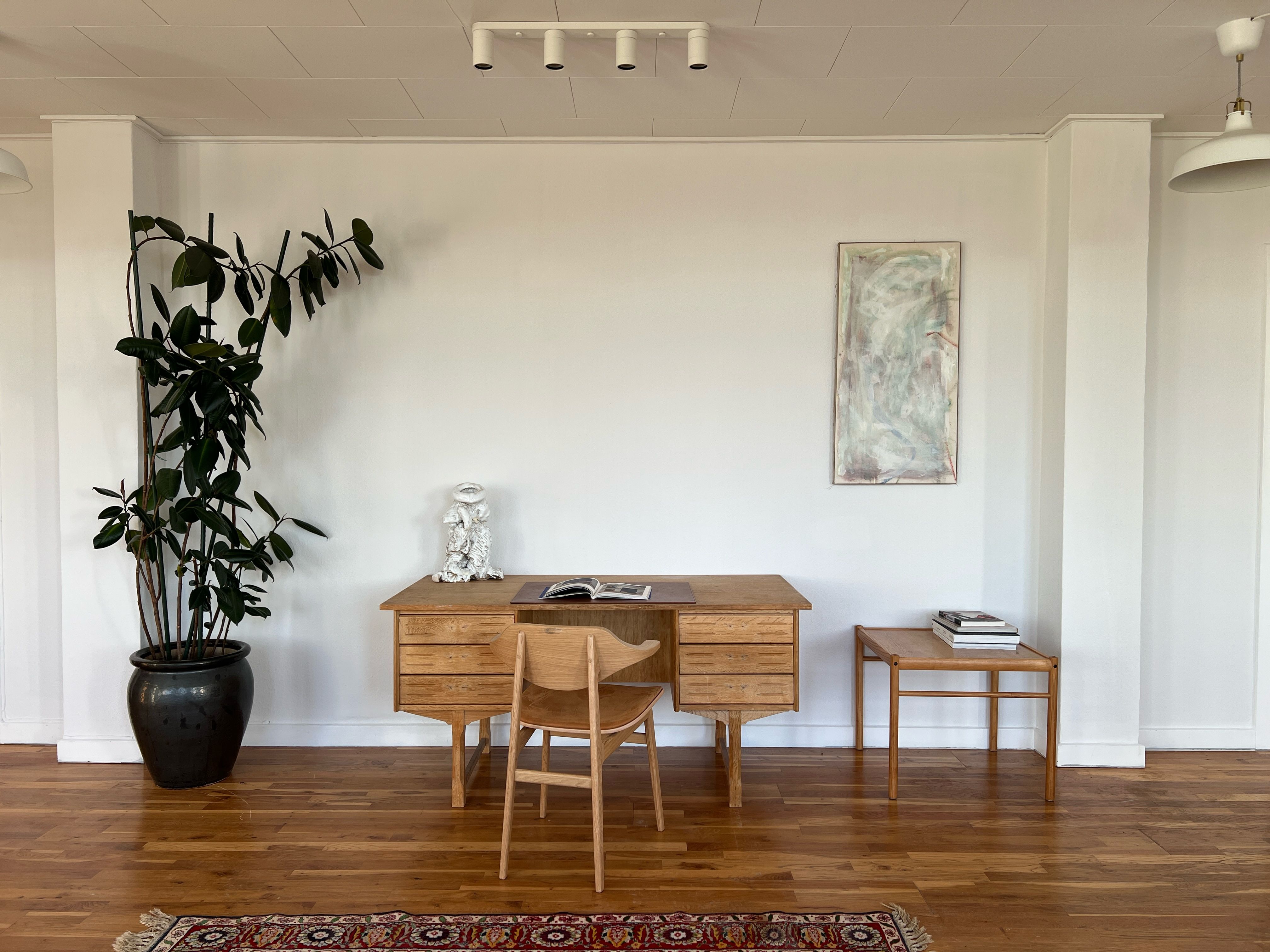 Kontor med trægulve, højloft og højt vindueslys. Bord og stol står på gulvet med plante og maleri på væggen.