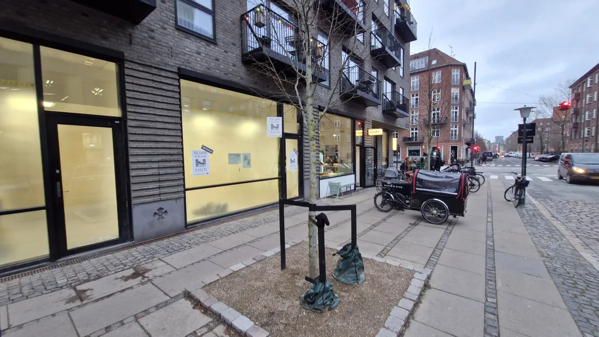 Fremlejemål med glasvinduer og døre på Ny Carlsberg Vej. To fiksterede cykler og en parkerede bil langs fortovet. Personer og bygninger på baggrunden.