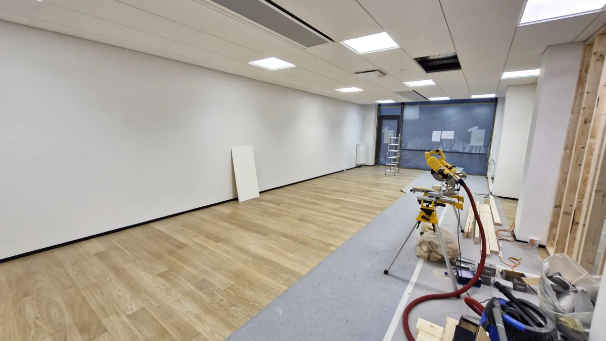 Et tomt rum med lyse lofter og hvide vægge, trægulv og gulvbelægning, samt diverse værktøj og materialer på gulvet og mod væggen.