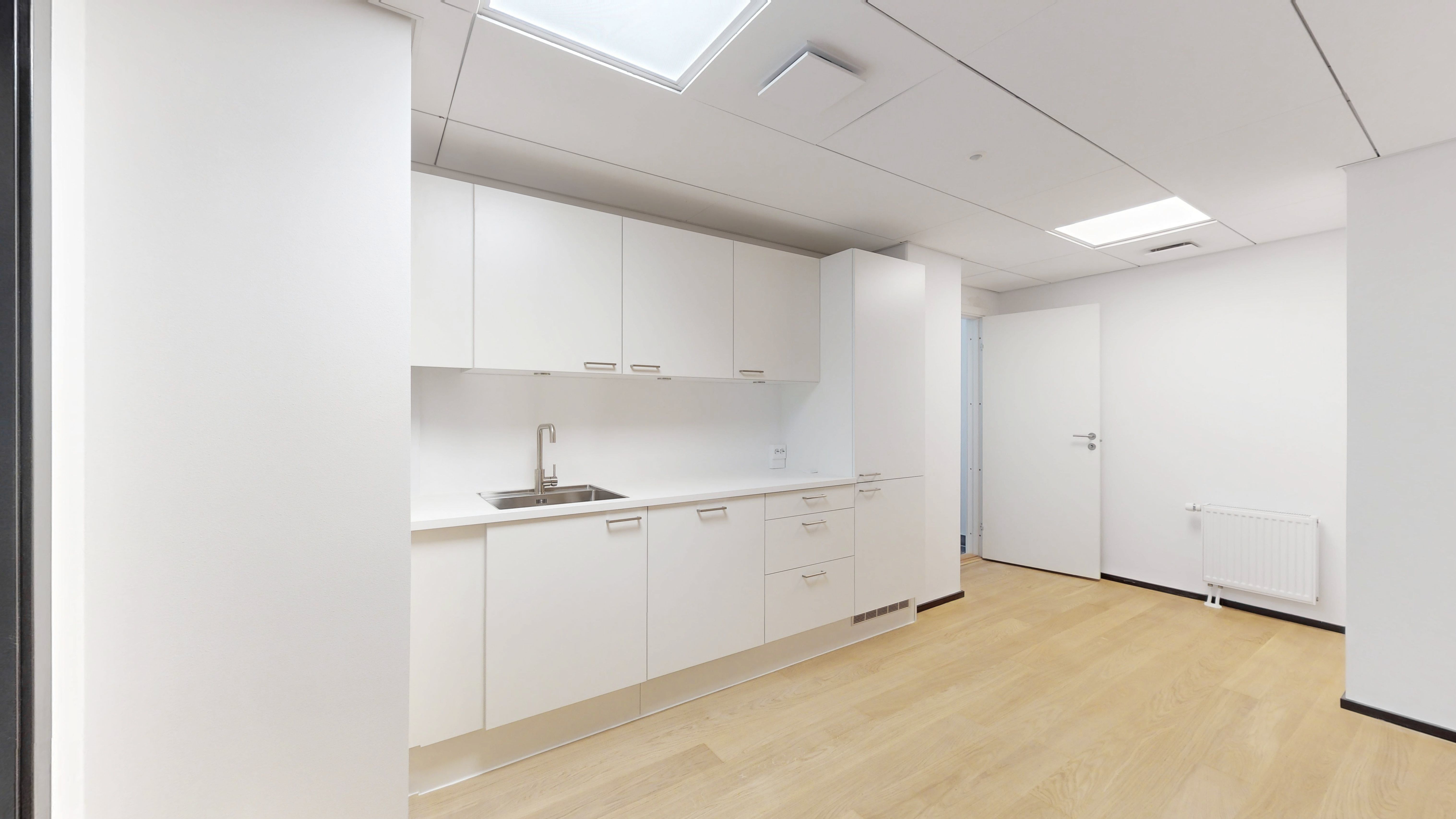 Det er et stort køkken med hvide vægge og lofter, lyse skygger og lyse skifertag, hvide skabe, en hvid radiator og en lysegrå gulv.