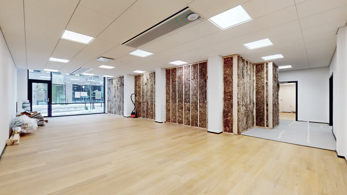 Udenfor en tom lejlighed i København er der en stor åbning i væggen, der er dækket med skifer. Loftet er hvidt og har flere runde loftslamper. Der er en søjle med hvid puds i midten af rummet.