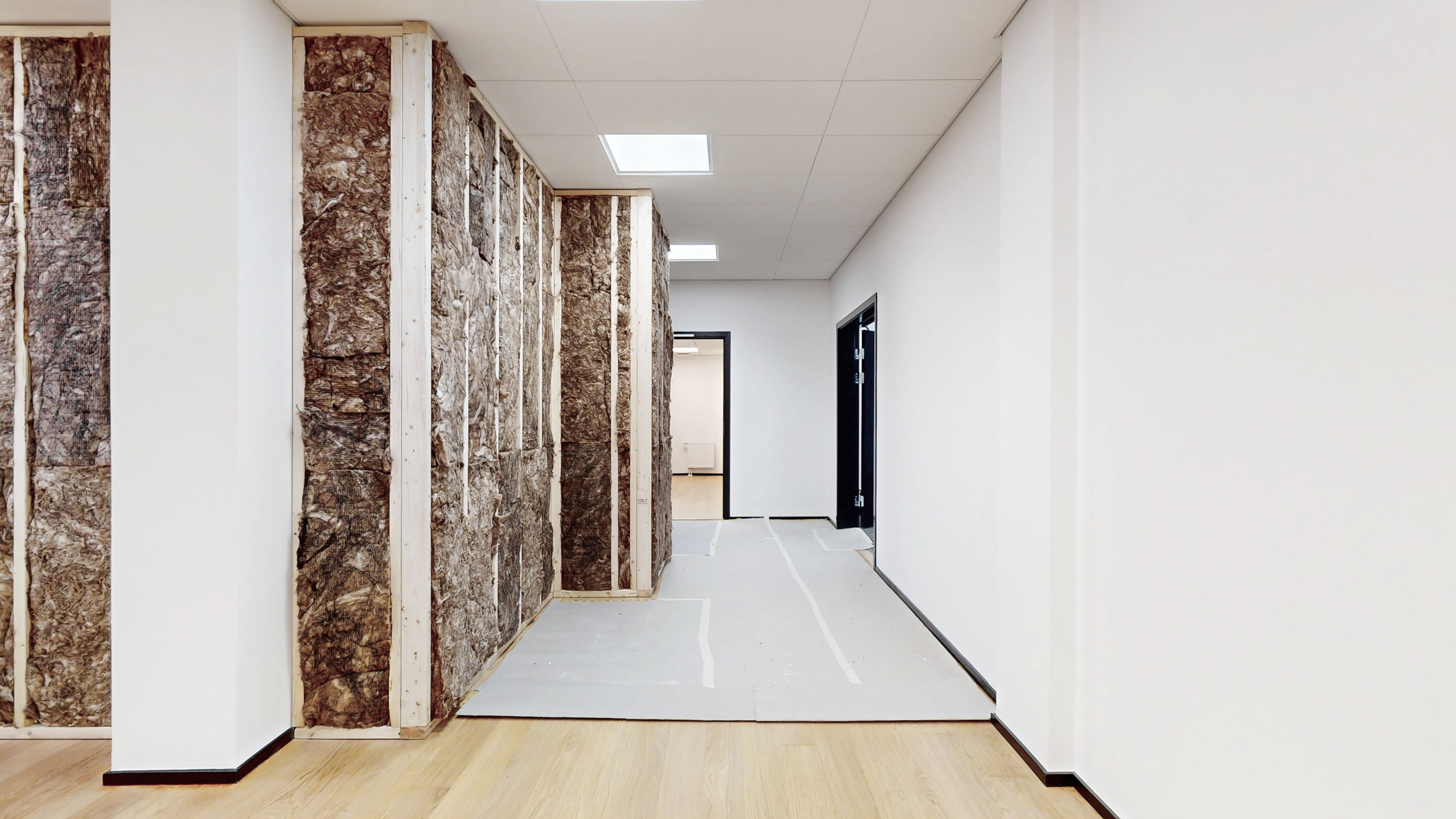 Hvid indvendig væg og gulv i et lejlighedslokal med skrå gulvbelægning og trægulv. Loftet er hvidt med flere hvide loftsskygger.