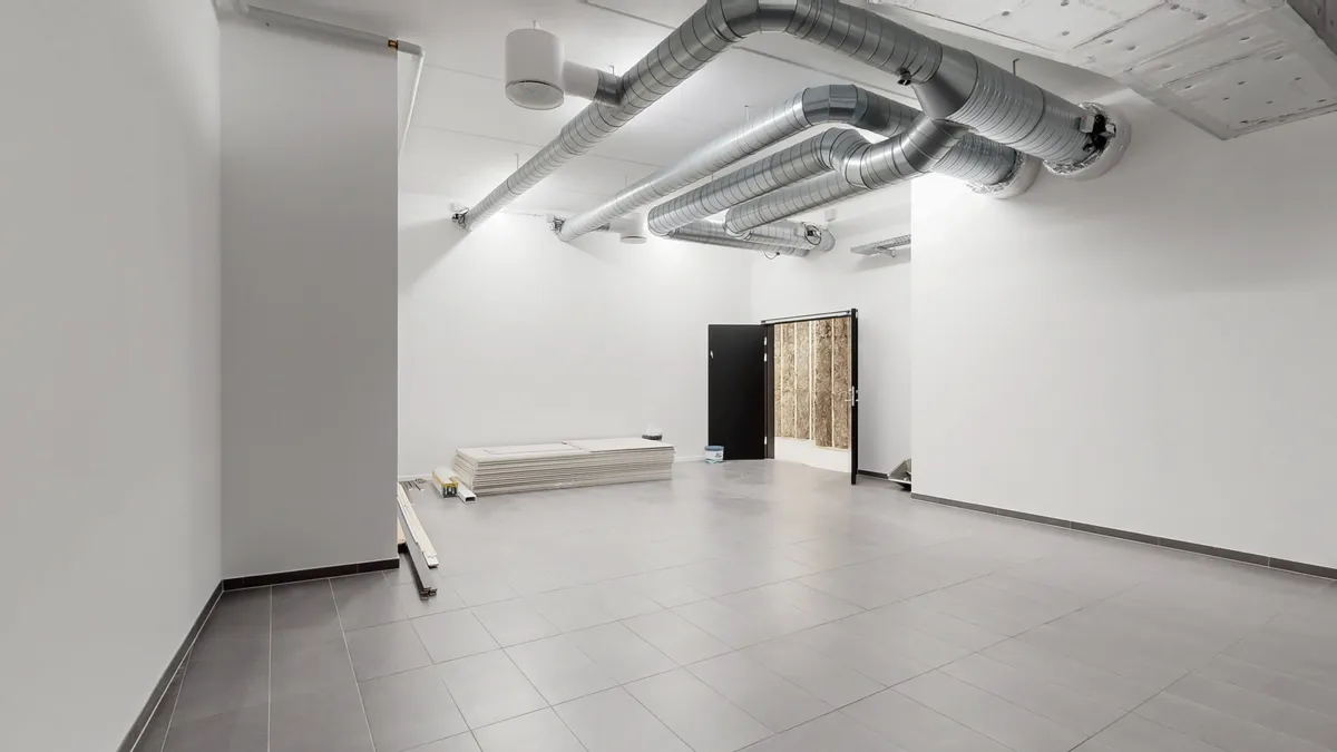 Ingen møbler og byggematerialer i det tomme rum med lysegule loftslampe. To vinduer med hvide rammer og sort dør i højre side.