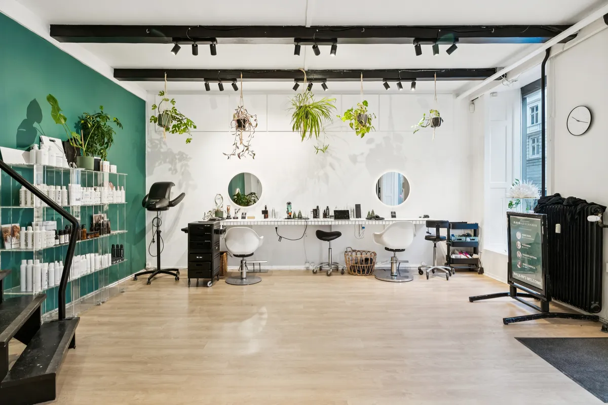 Lyst og moderne frisørsalon med hvide vægge, lyse gulve og grønne vægplante. Salonen er udstyret med flere barberstole, spejle og et stort produkthylde