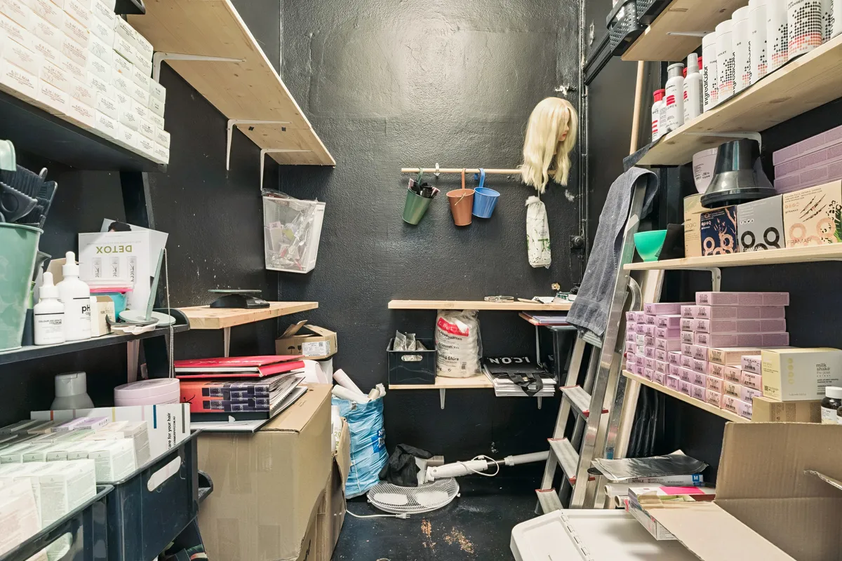 Butikslokale med opbevaringsskabe og hylder med varer og emballage, kassevogn og stige samt tøj og tøjstander og en pupper i et hjørne