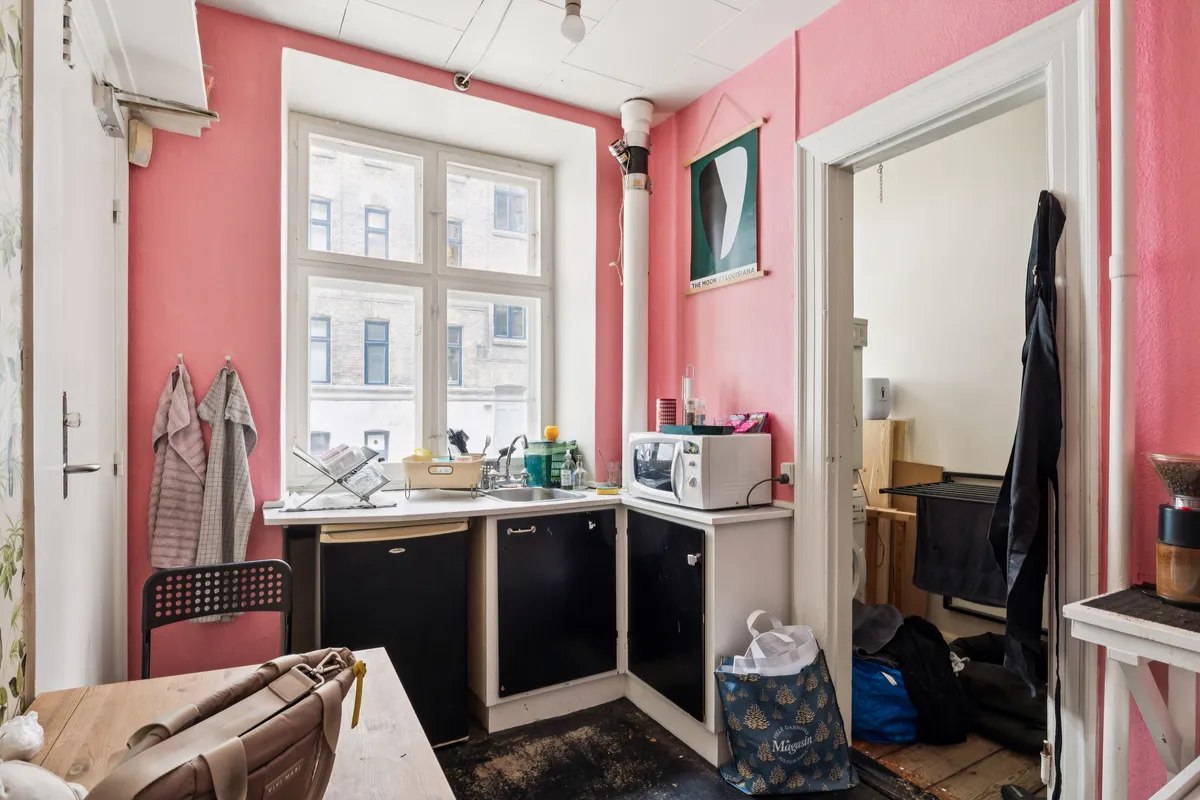 Alt-tekst: Et køkken med høje, lyse pink vægge og møbler i sort og hvidt. Der er en mikrobølgeovn, køkkenredskaber og to hængeklæder.