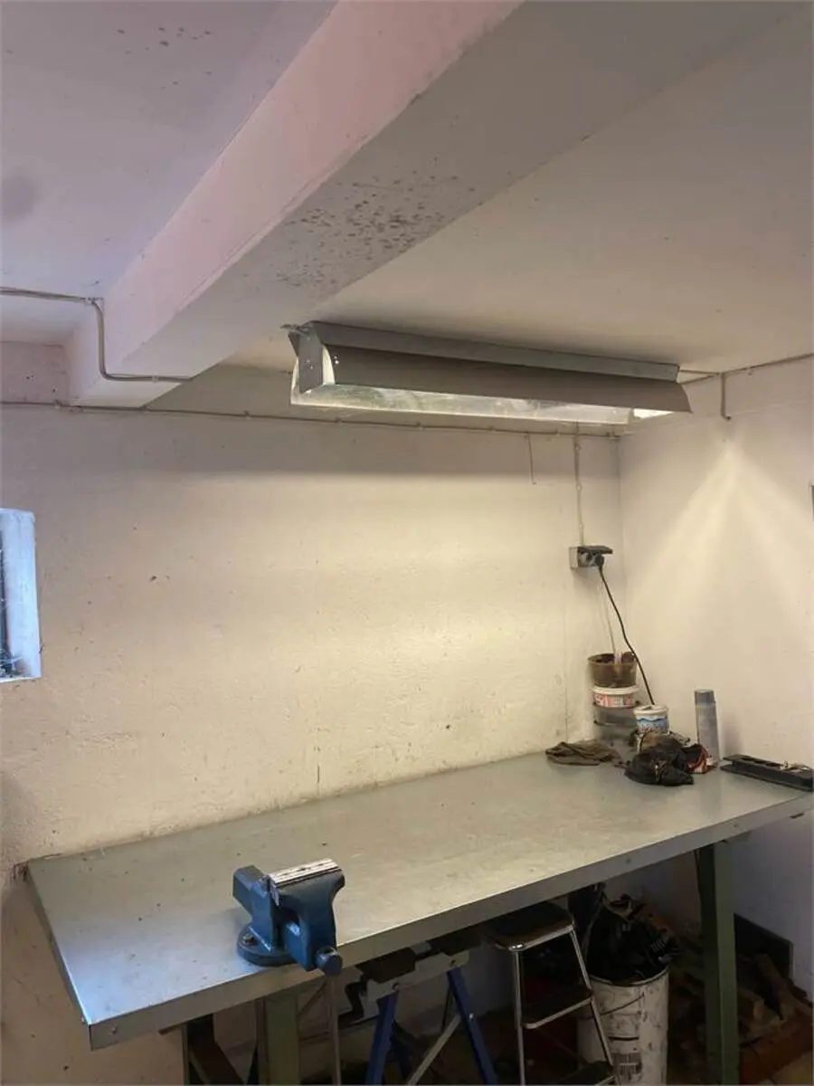 Produktionsværksted med metalbord, svejsemaskine og værktøjskurv. Forskelligt værktøj samt materialer er placeret på væggen. Loftslampe og væggen er snavset.