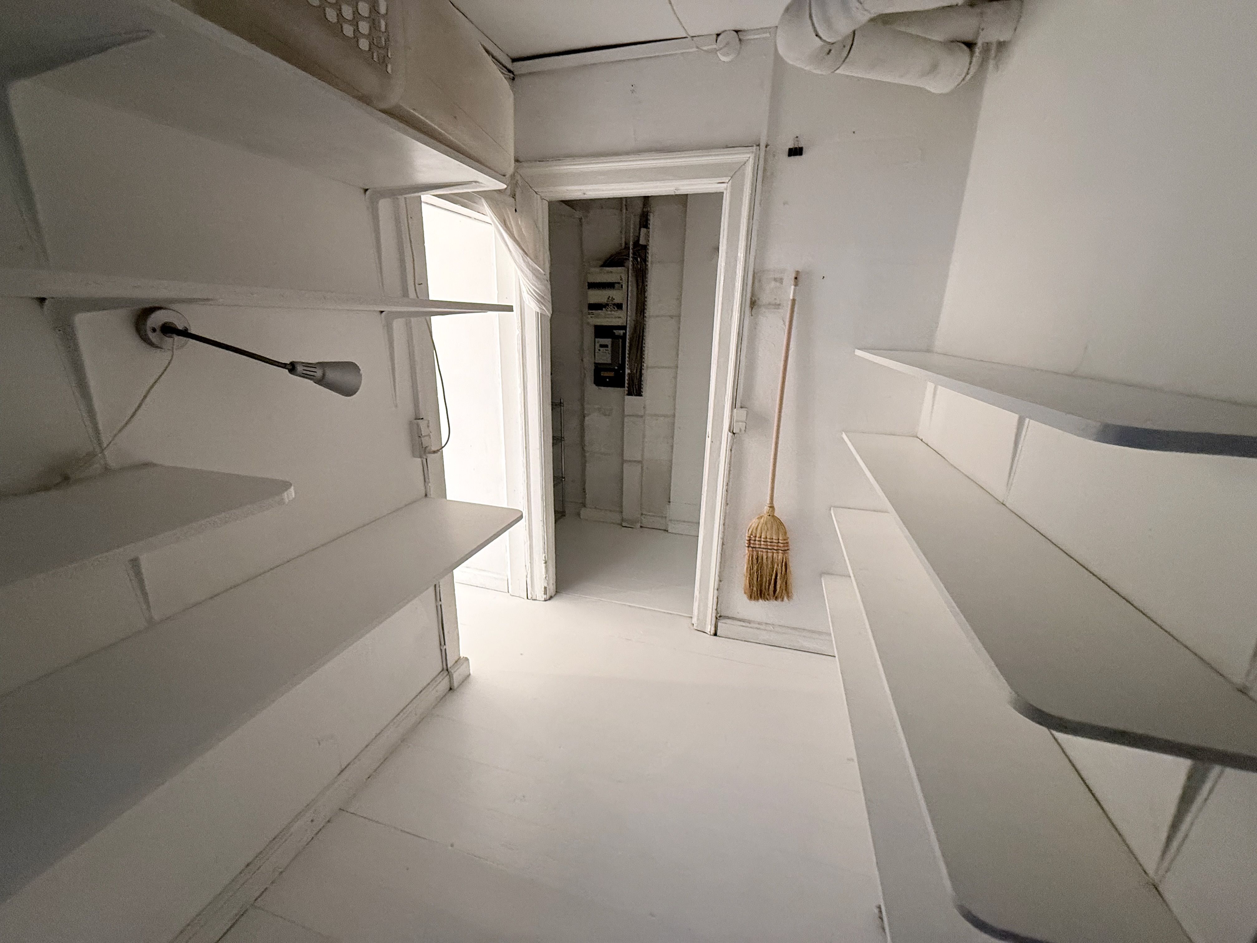 Lokaler med hvide vægge og højloft samt lysegule gulvbelægning, to rum med højloft, en åben dør, to bjælker med hvide hylder og et hængende lampe.