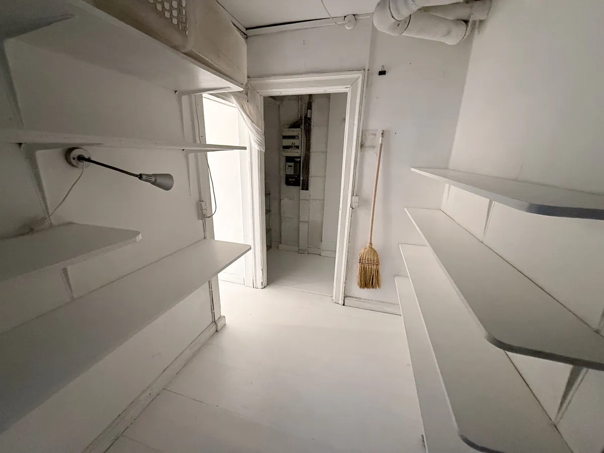 Lokaler med hvide vægge og højloft samt lysegule gulvbelægning, to rum med højloft, en åben dør, to bjælker med hvide hylder og et hængende lampe.