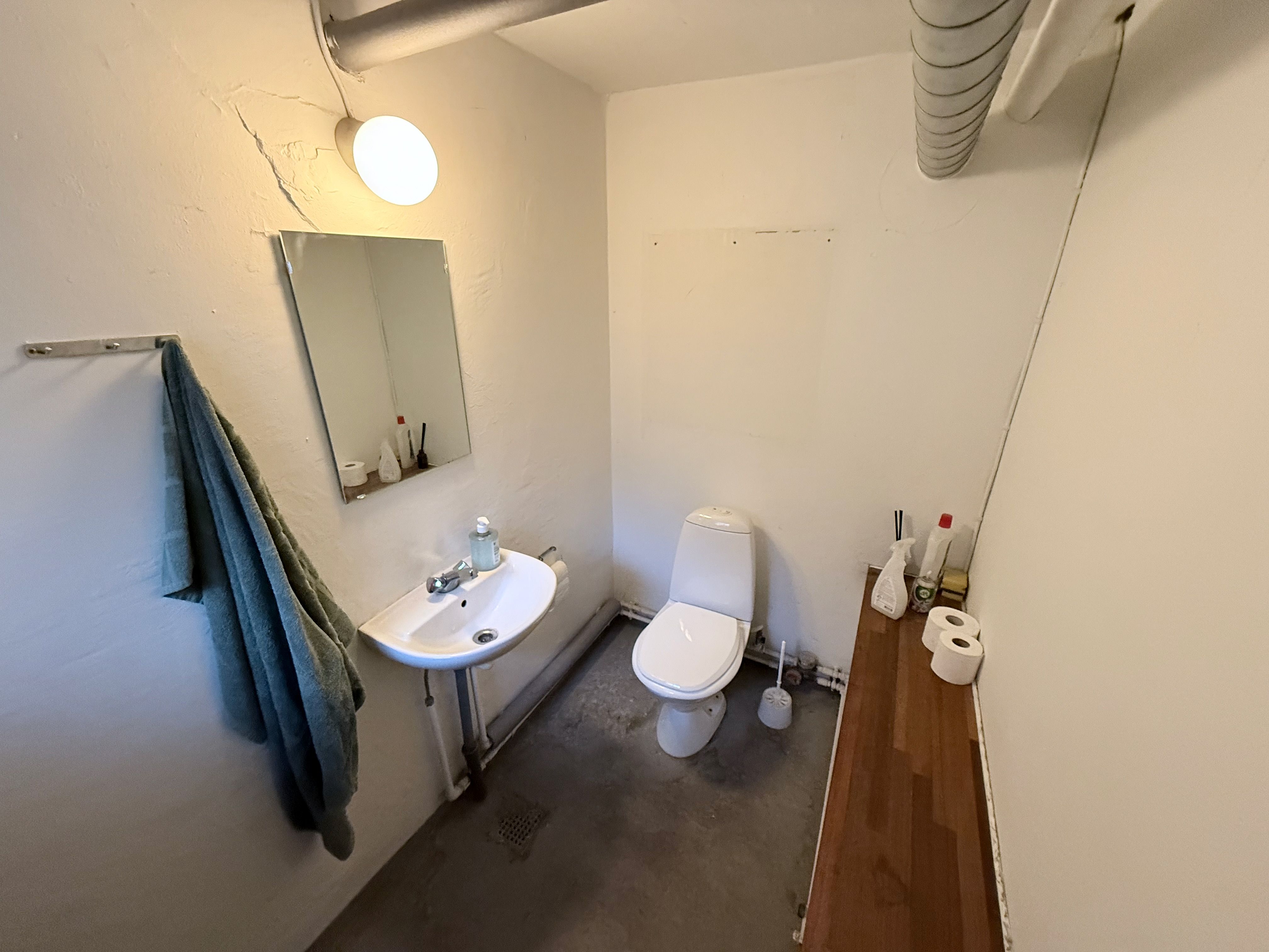 Billede af et lille toiletrum med toilet, håndvask, spejl og to ruller toiletpapir. Rummet er rent og velplejet med hvidt vægpanel.