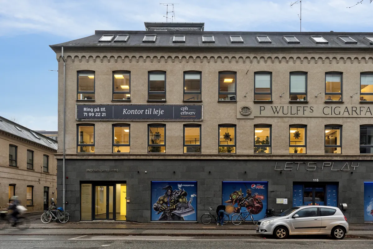 Bygning med facade 115 og reklame for Predator og P. Wulffs Cigarfabrik. Lejlighed 113 med stort vindue og lejetegn. Cykler og bil parkeret foran.