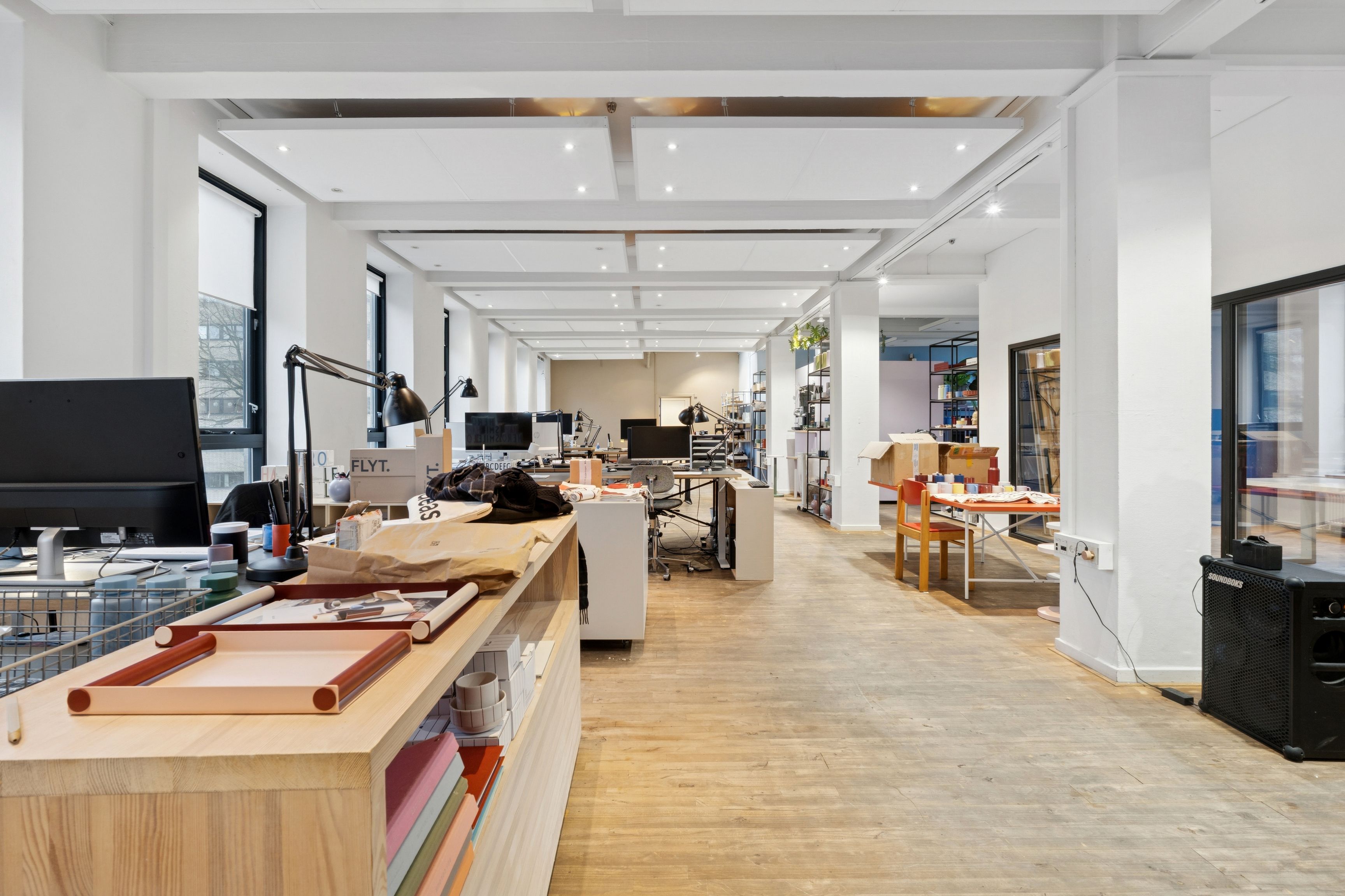 Kontor med computere, stole, skriveborde, lys, og møbler i en stor rummelig stue med højt loft og trægulve