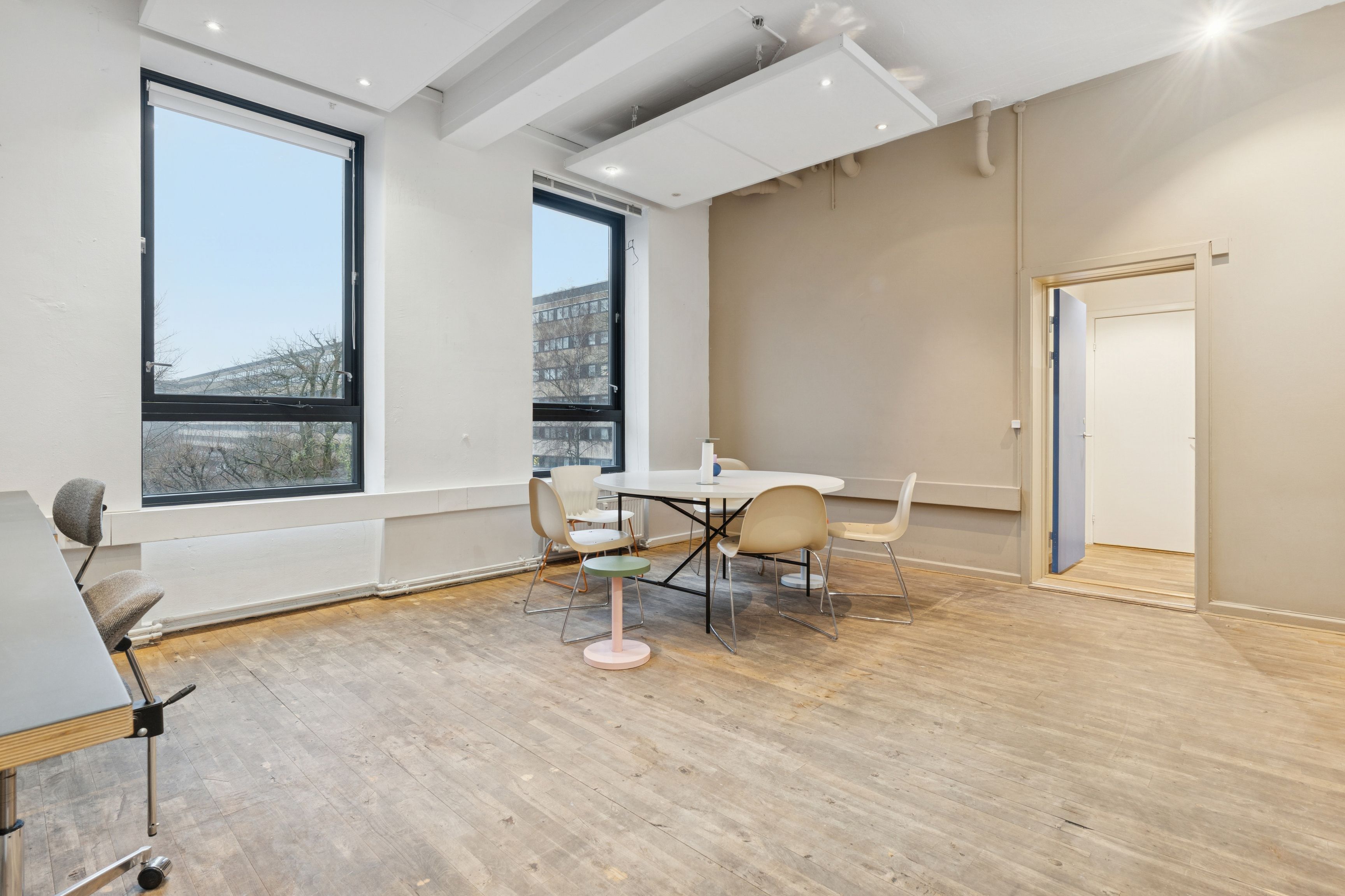 En stor og lyse kontorlokal med trægulve, hvide vægge og vinduer med udsigt til bygninger. Fire stole og et rundt bord er placeret i midten af rummet.