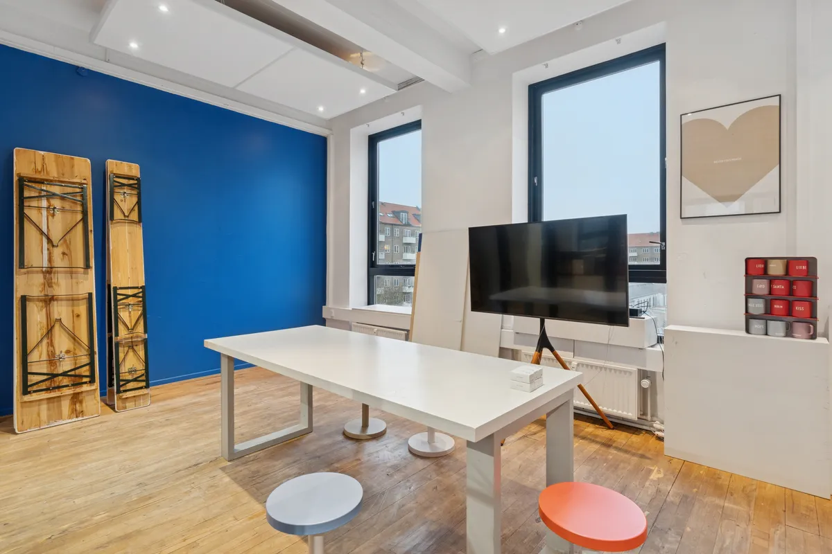 Billede af et stort kontor med lysegul væg og møbler i hvidt og lysebrunt, to stole, højt tv og loftsventilatorer samt et træbord med små bokse