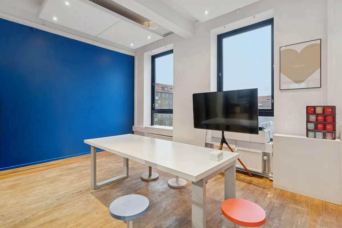 En stor rummelig stue med hvide vægge og gulv samt en stor blå væg og en stor sort tv-skærm samt to stole og en lang hvid bord