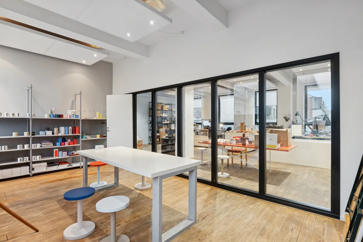 Værelse med højt loft, lyse gulve og store vinduer med udsigter til byen. Værelse med bord og stole samt hylder med diverse genstande.